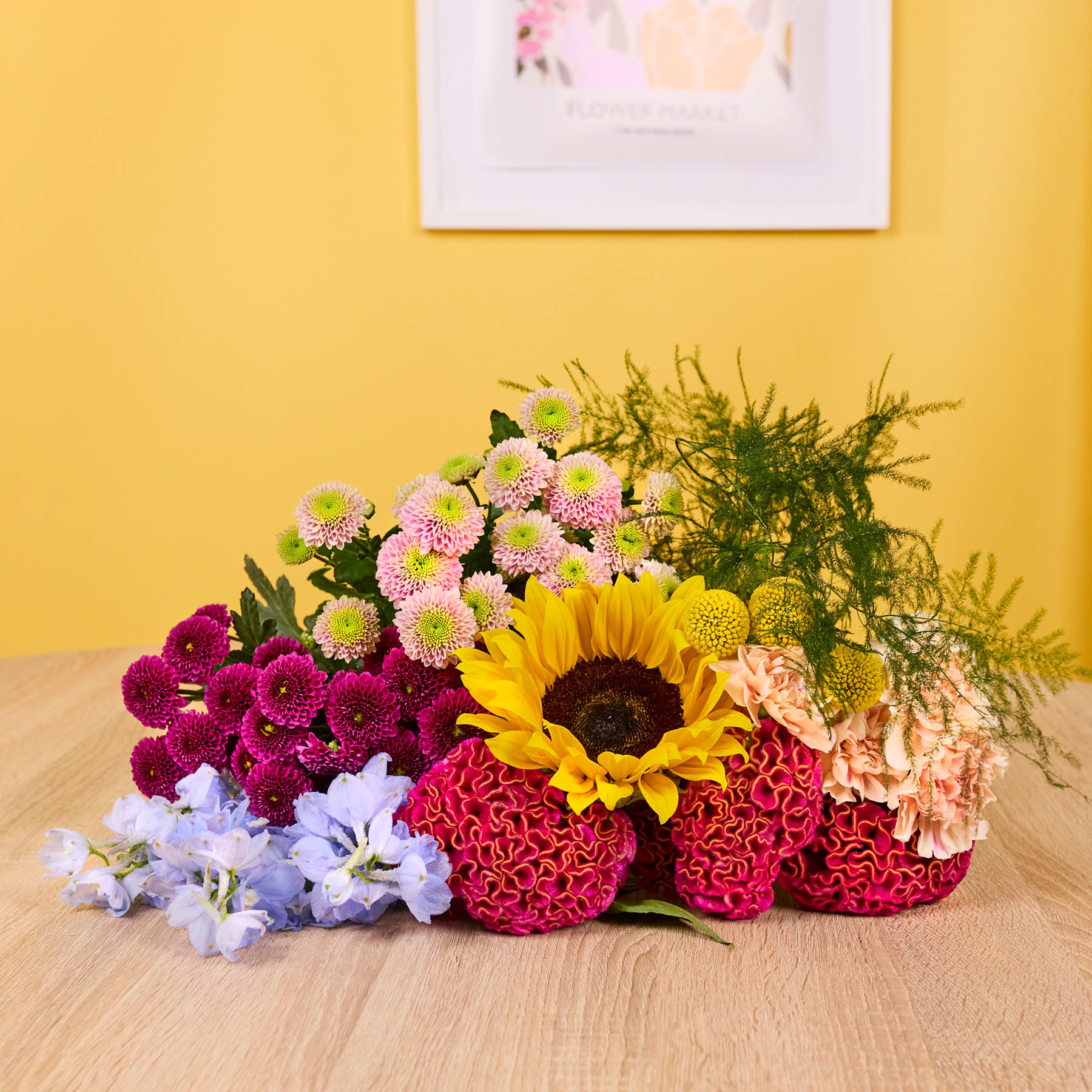 Ein farbenfroher Strauß bunter Blumen, darunter Sonnenblumen, Chrysanthemen und Nelken, steht auf einer Holzoberfläche vor einer gelben Wand mit einem gerahmten Kunstwerk.