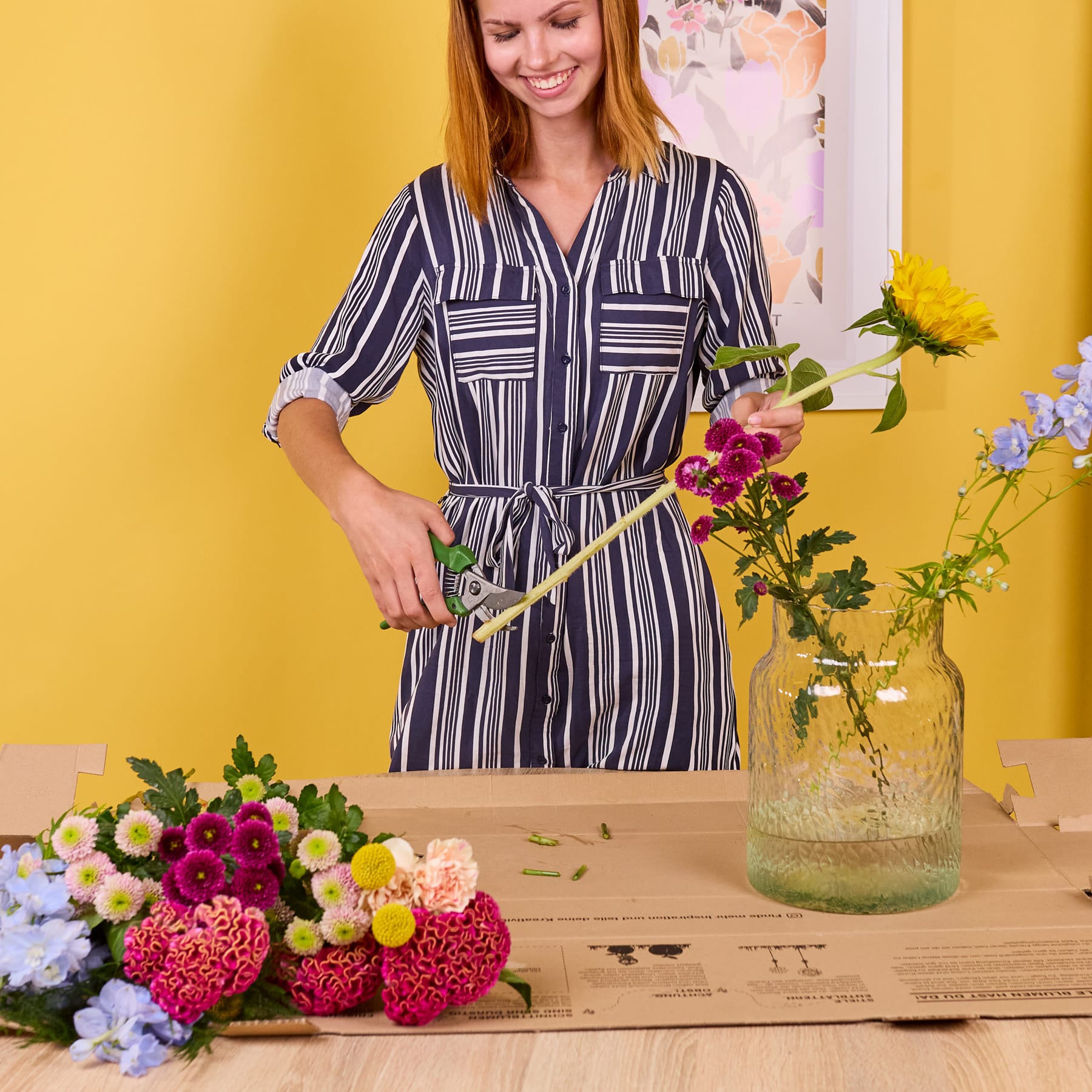Eine junge Frau in einem gestreiften Kleid steht vor einer gelben Wand, umgeben von einem Strauß leuchtender Blumen auf einer Holzkiste.