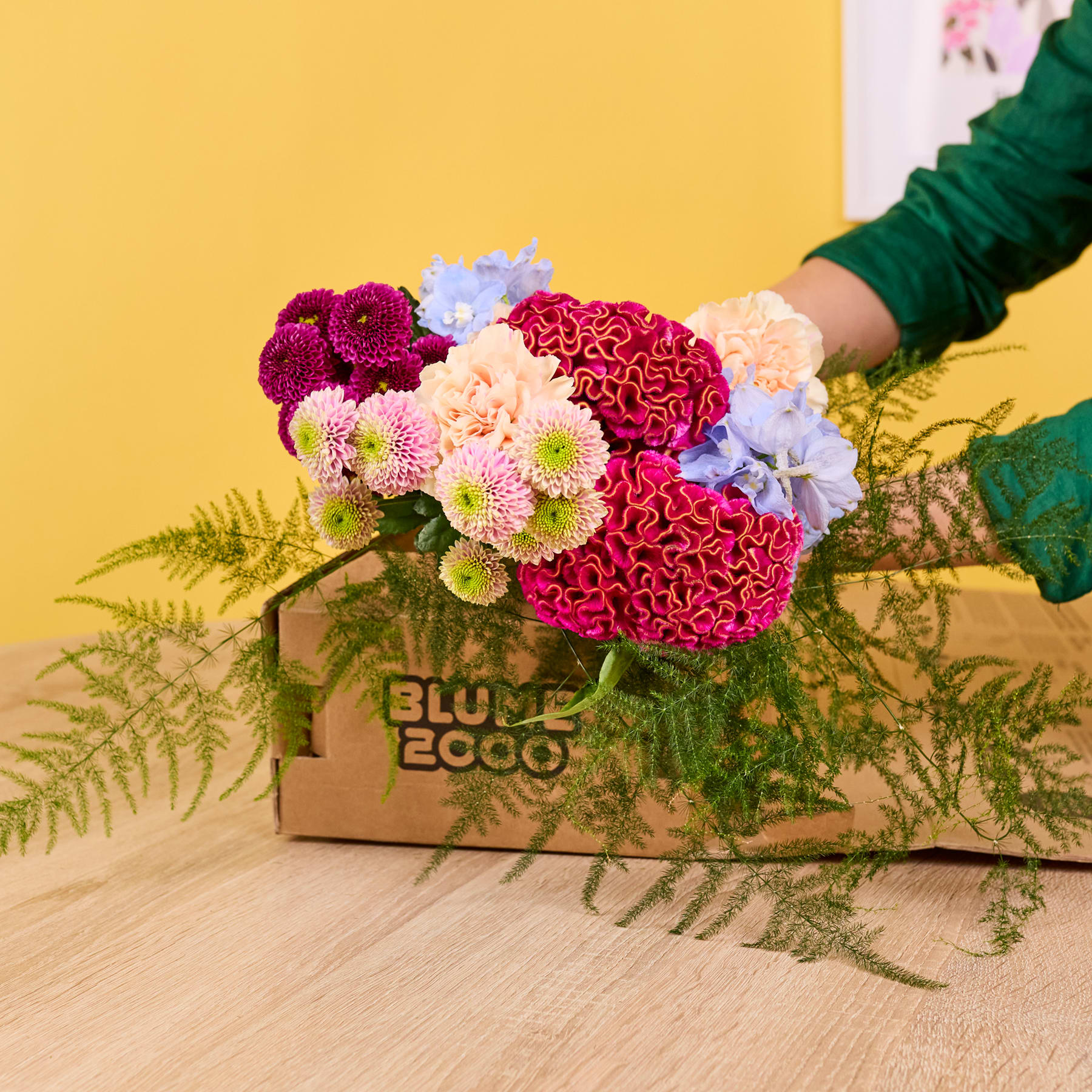 Ein lebendiger Strauß bunter Blumen, darunter rote und rosa Dahlien, eingebettet in eine Holzkiste vor einem leuchtend gelben Hintergrund.
