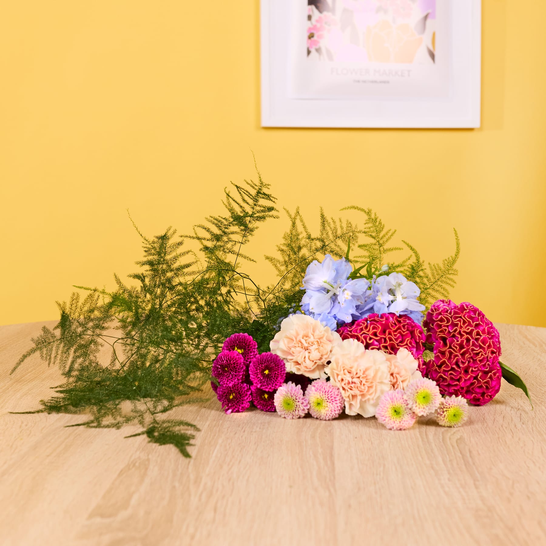 Ein leuchtender Strauß bunter Blumen, darunter rosa Dahlien, rote Chrysanthemen und blaue Hortensien, steht auf einer Holzoberfläche vor einer gelben Wand mit einem gerahmten Kunstwerk.