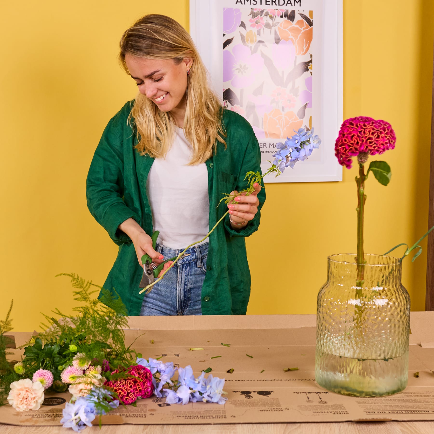 Eine junge Frau in einer grünen Jacke steht vor einer gelben Wand, umgeben von verschiedenen Blumen und Pflanzen auf einem Holztisch.