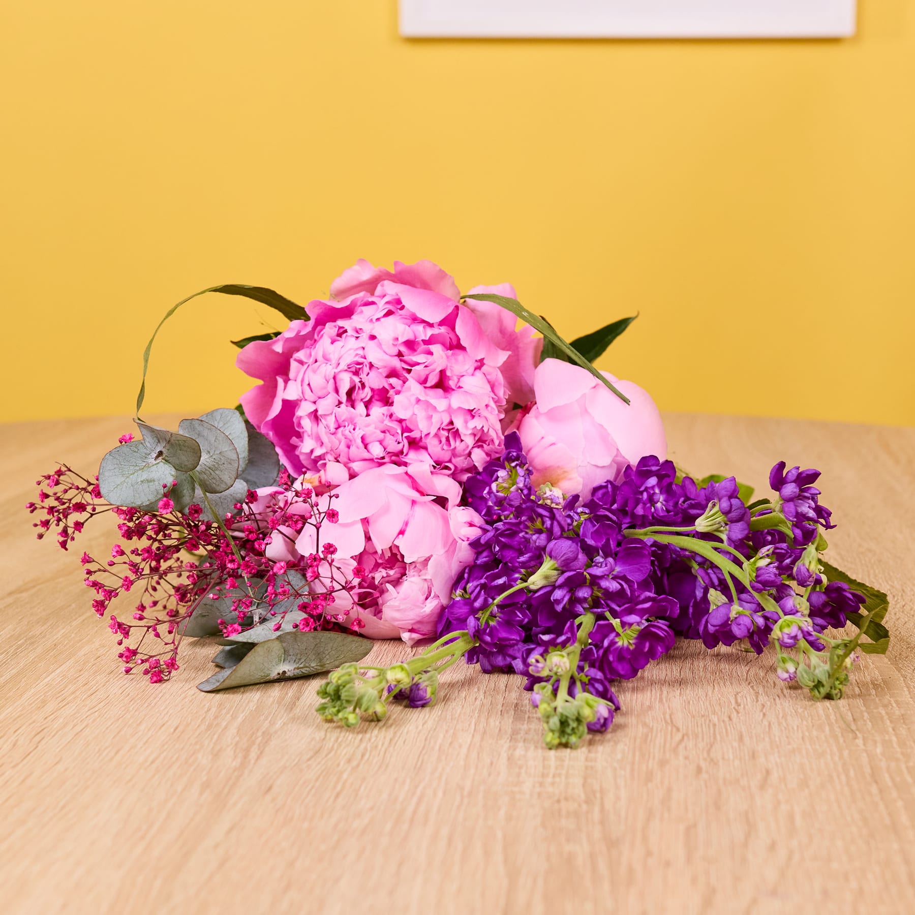 Ein farbenfroher Strauß aus rosa Pfingstrosen, violetten Schwertlilien und Grünpflanzen steht auf einer Holzoberfläche vor einem leuchtend gelben Hintergrund.