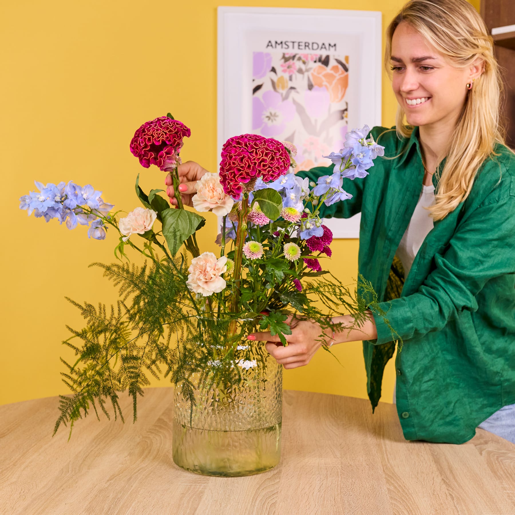 Eine lächelnde Frau in einer grünen Jacke arrangiert einen farbenfrohen Blumenstrauß auf einem Holztisch, im Hintergrund ist ein buntes Poster von Amsterdam zu sehen.