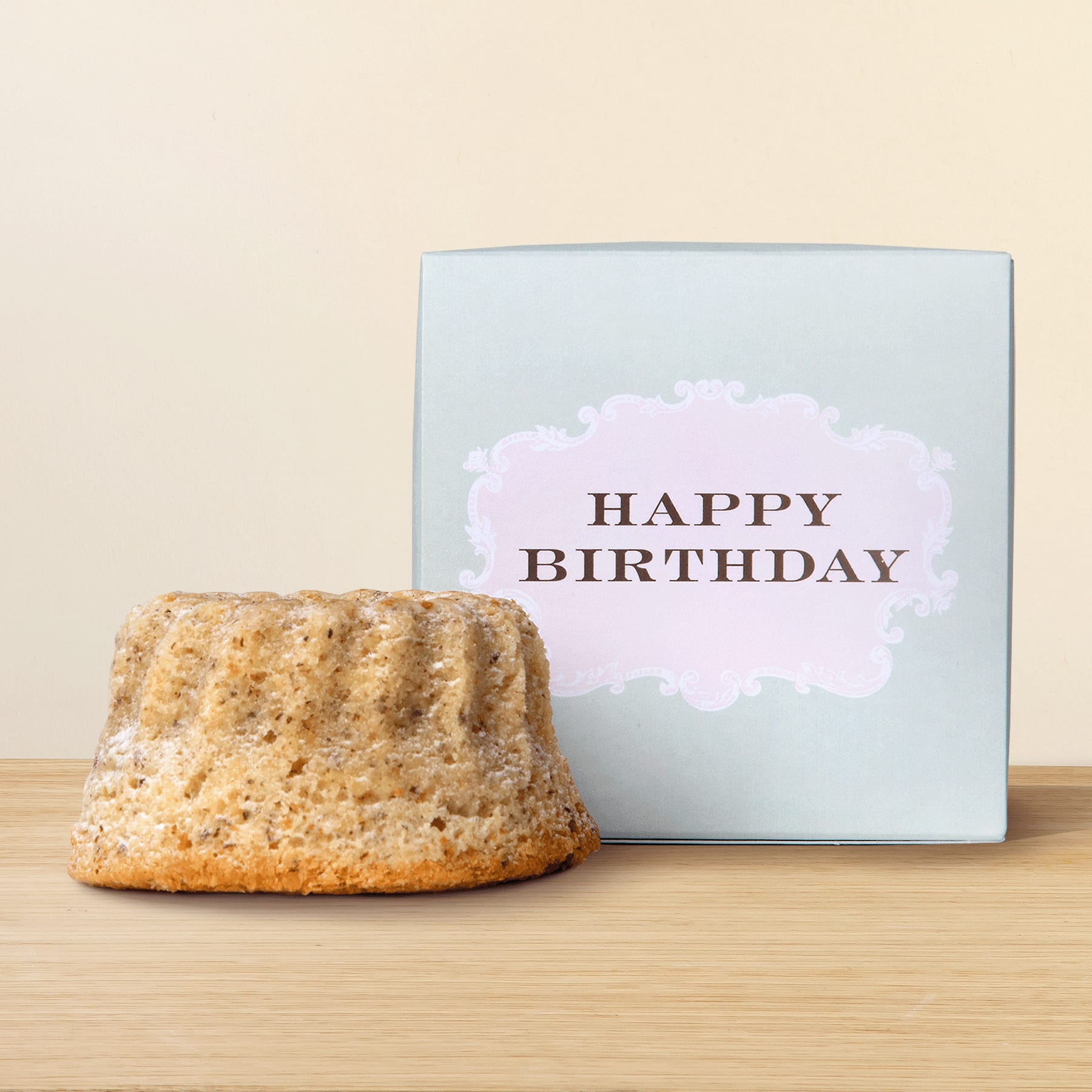 Auf einer Holzoberfläche steht ein heller Kuchen oder Gebäck, daneben eine hellblaue quadratische Schachtel mit der Aufschrift „HAPPY BIRTHDAY“.