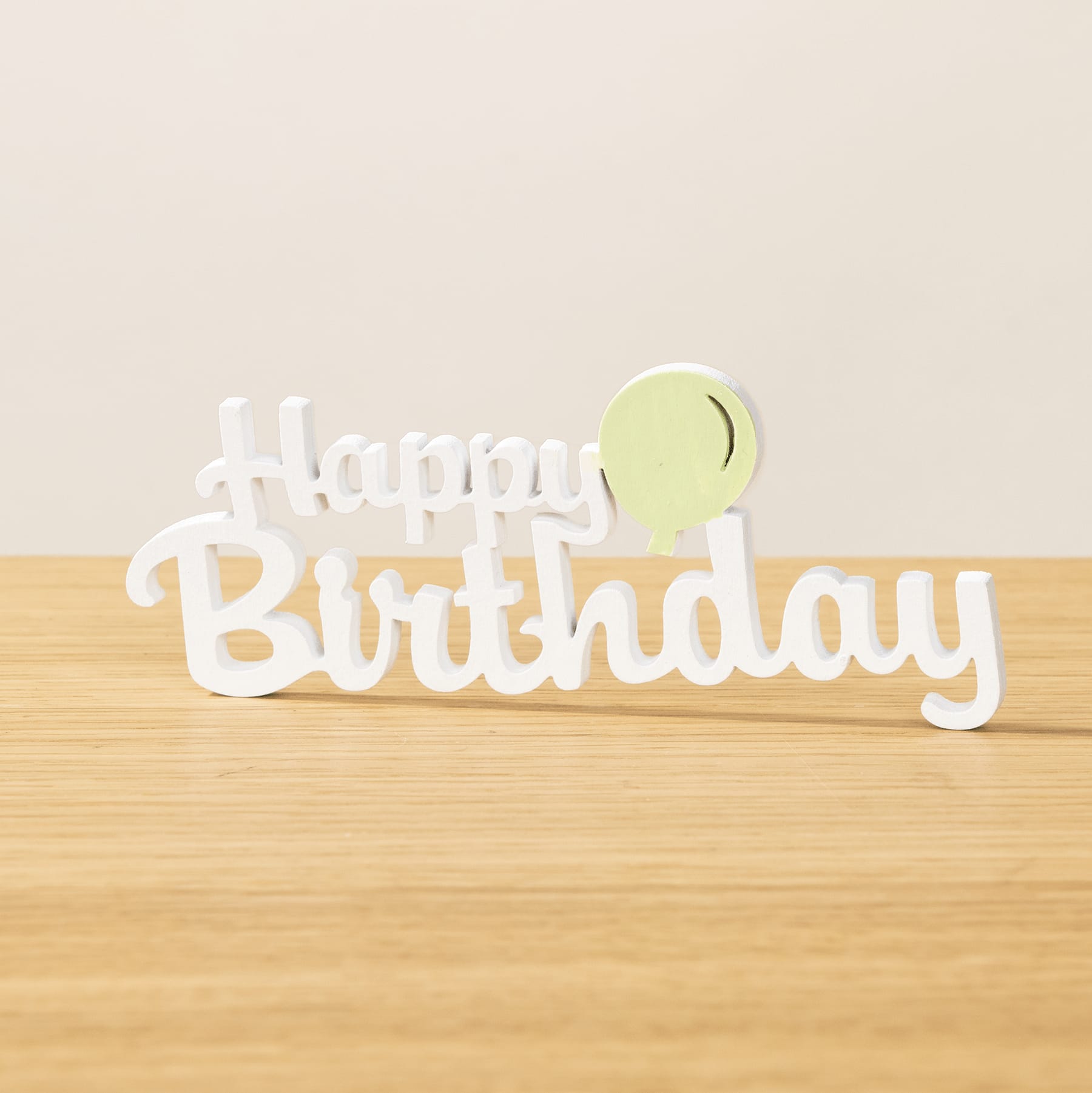 Ein weißes „Happy Birthday“-Schild steht auf einer Holzoberfläche vor einem schlichten, hellen Hintergrund.