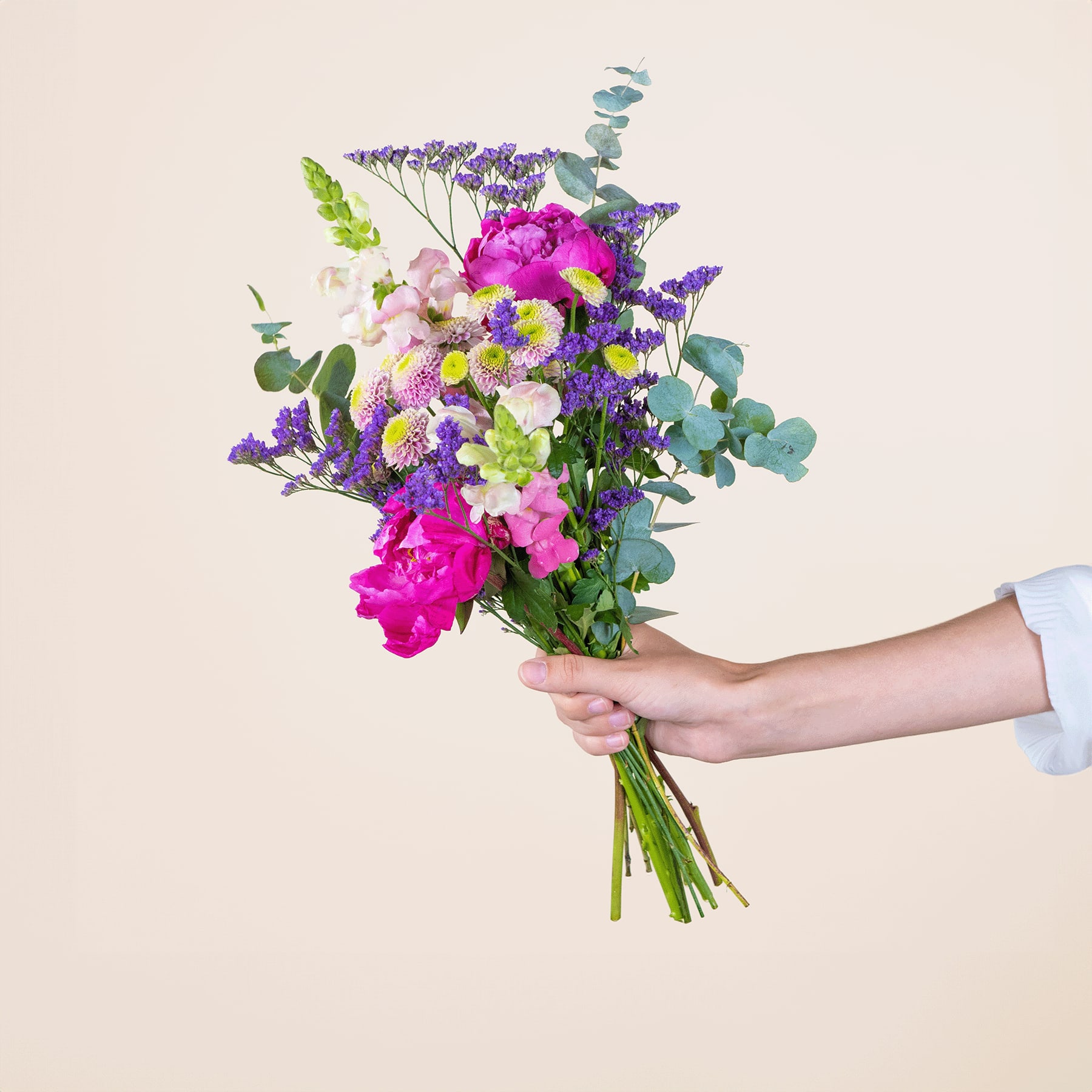 Ein leuchtender Strauß bunter Blumen, darunter rosa Pfingstrosen, lila Lavendel und grünes Laub, wird vor einem schlichten beigen Hintergrund in der Hand einer Person gehalten.