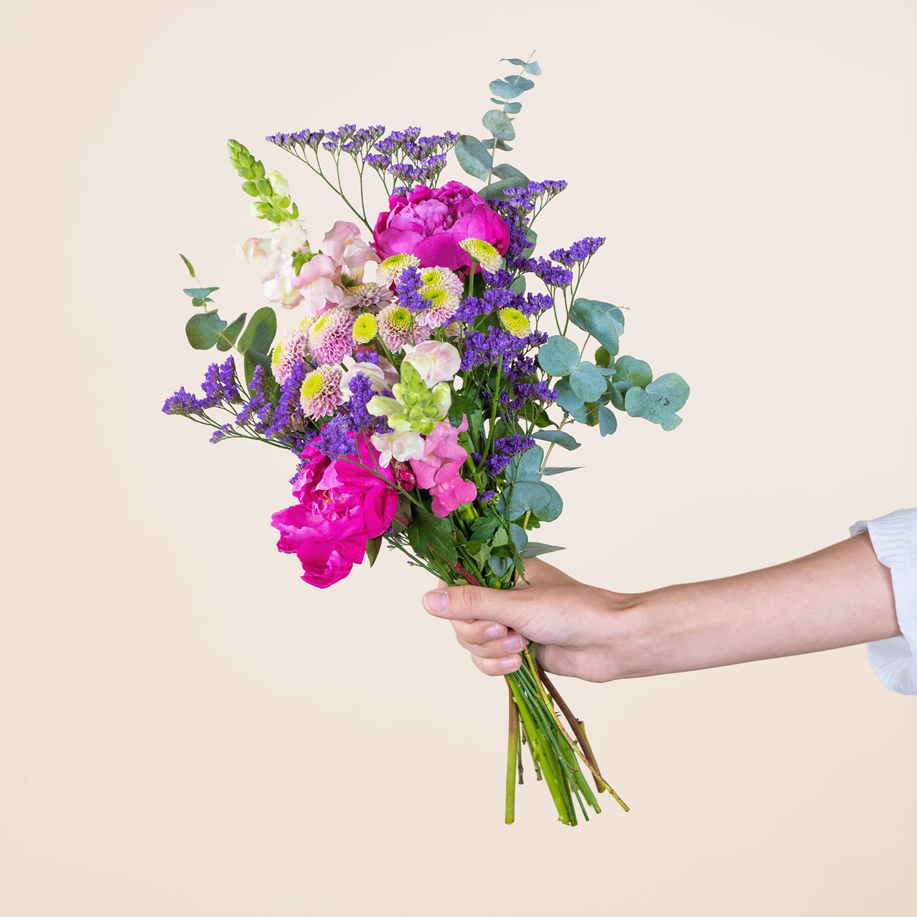 Ein leuchtender Strauß bunter Blumen, darunter rosa Pfingstrosen, lila Lavendel und grünes Laub, wird vor einem schlichten, hellen Hintergrund in den Händen einer Person gehalten.