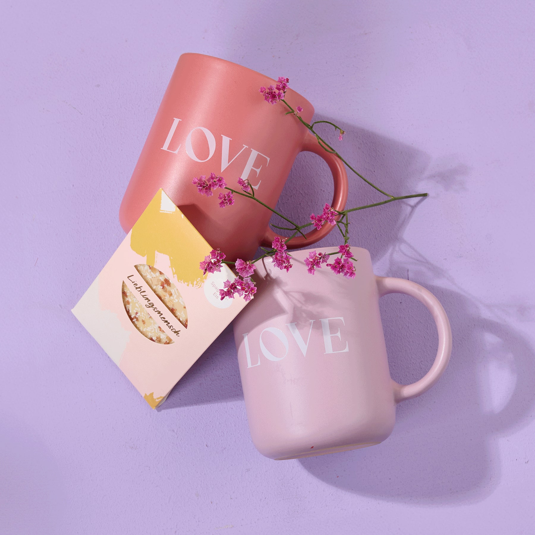 Vor einem zarten lavendelfarbenen Hintergrund stehen zwei Tassen mit dem Aufdruck „LOVE“ und einem kleinen Strauß lila Blumen.