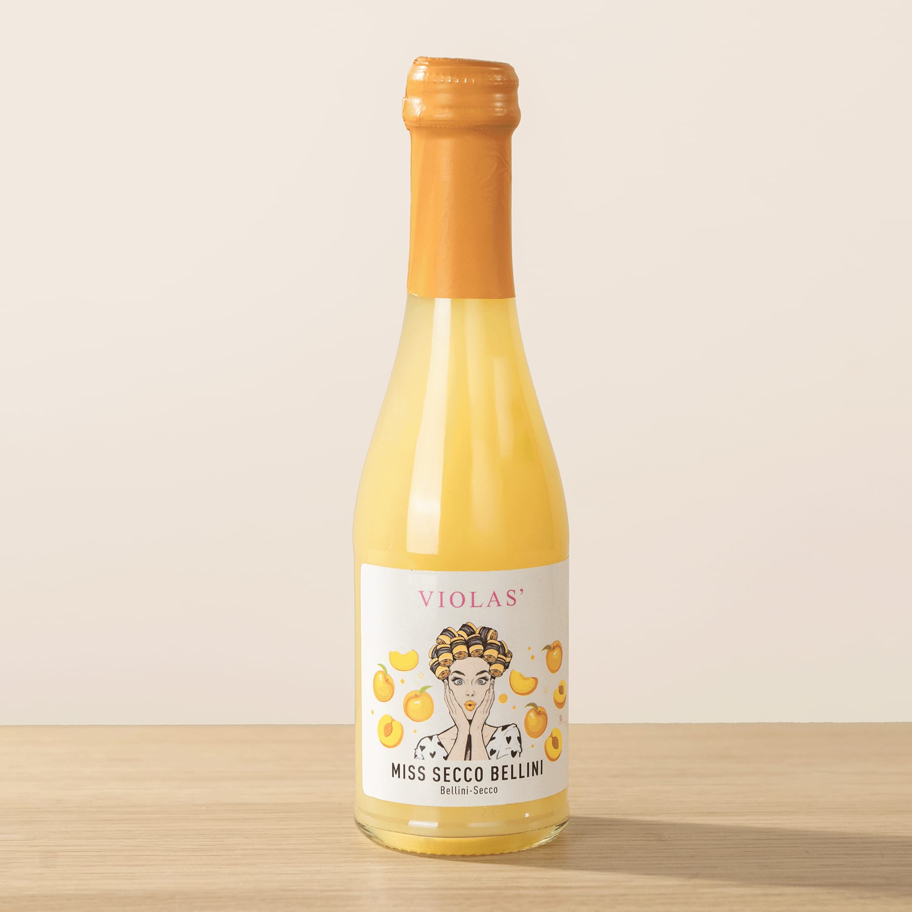 Eine gelbe Glasflasche mit orangefarbenem Verschluss und einem Etikett mit der stilisierten Illustration eines Frauengesichts und dem Text „VIOLAS‘ MISS SECCO BELLINI“ steht auf einer Holzoberfläche vor einem hellen Hintergrund.