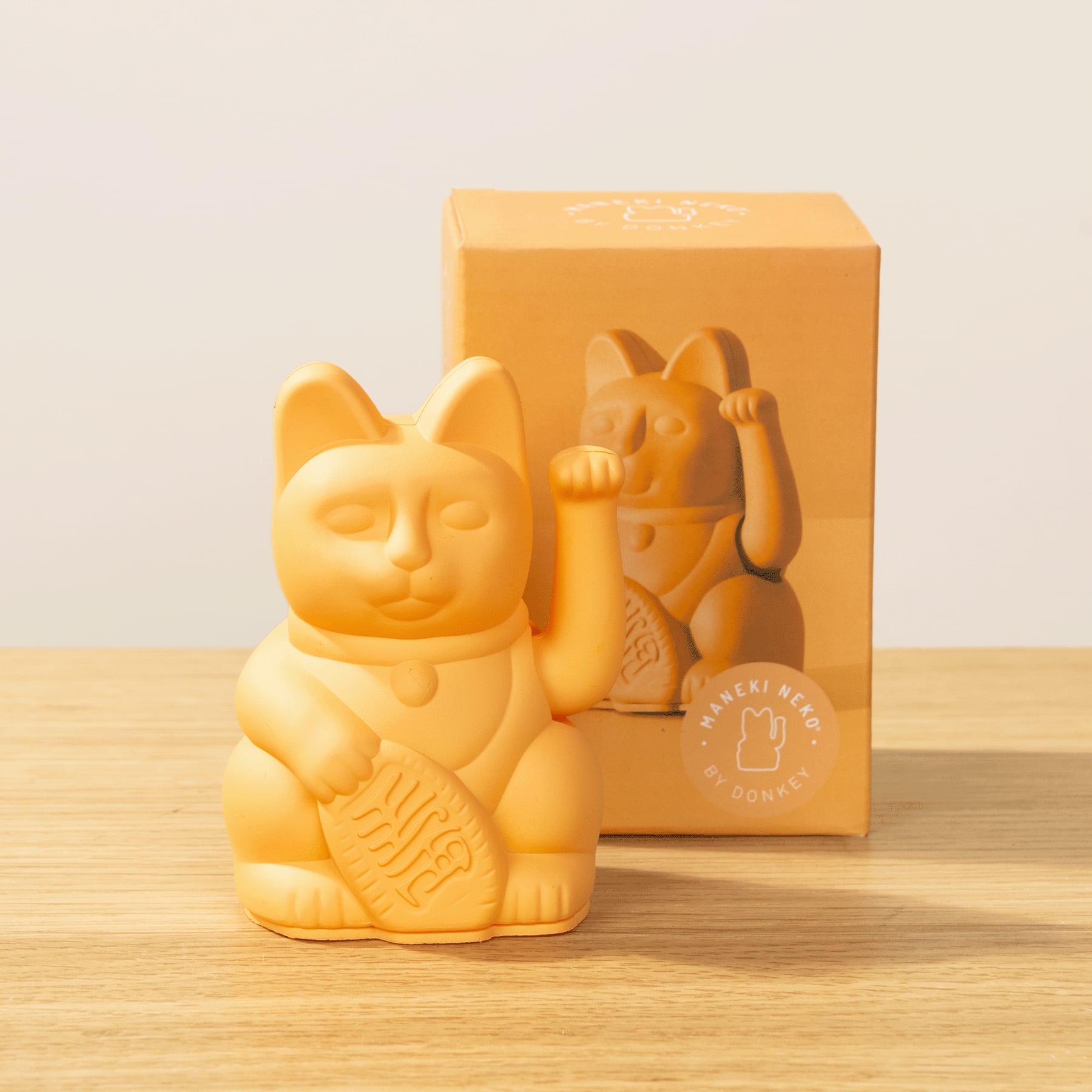 Eine gelbe Keramikfigur eines katzenartigen Wesens, bekannt als „Maneki-neko“ oder „Glückskatze“, sitzt auf einer Holzoberfläche mit einer gelben Schachtel im Hintergrund.