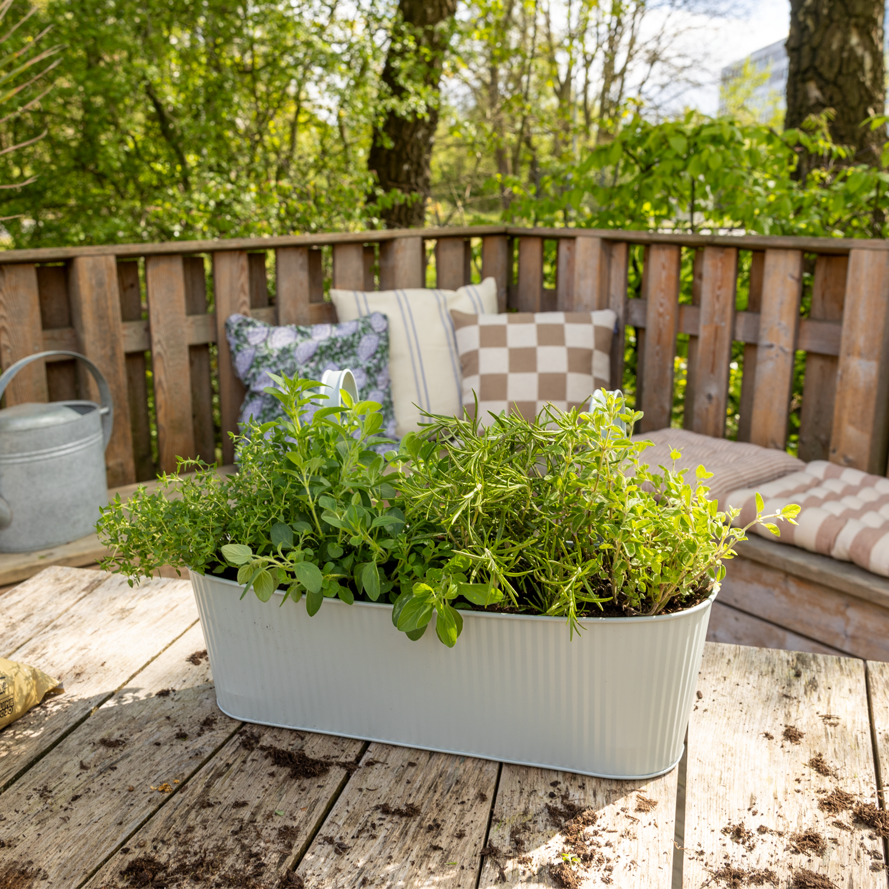 Eine Holzterrasse mit einem Pflanzkasten voller üppiger, grüner Kräuter und Pflanzen, umgeben von einem Holzzaun und einem grünen, bewaldeten Hintergrund.