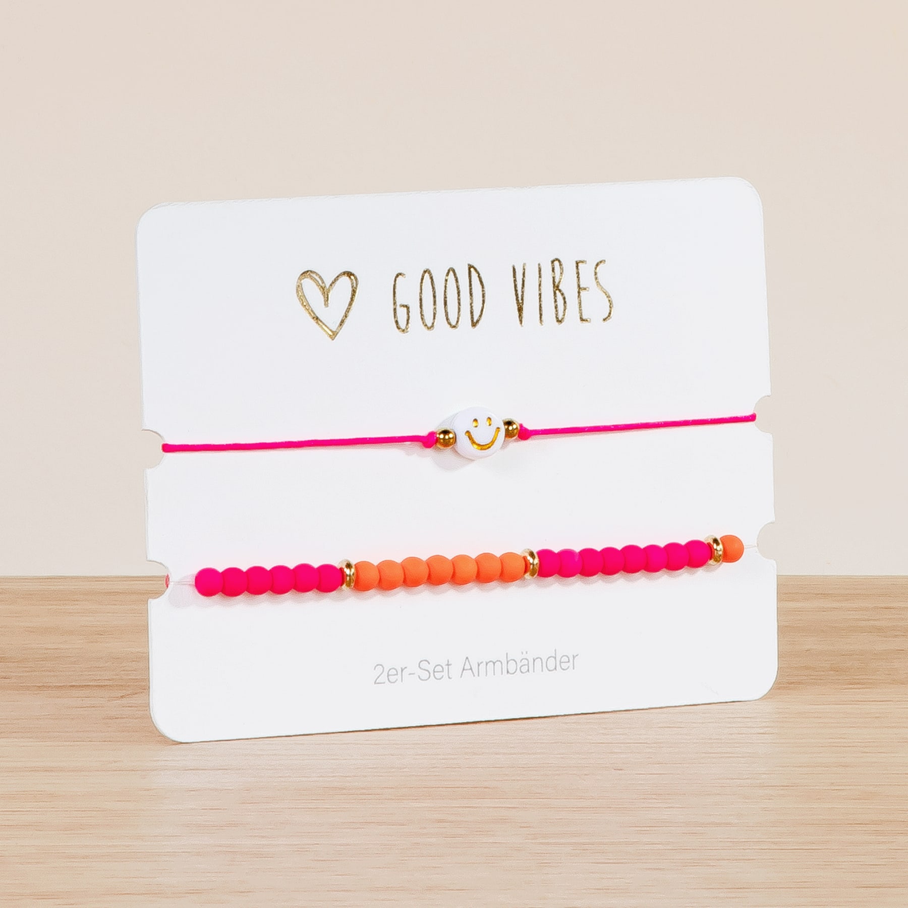 Das Bild zeigt ein Set bunter Perlenarmbänder auf einer weißen Karte mit dem goldenen Schriftzug „GOOD VIBES“. Die Armbänder sind optisch ansprechend angeordnet, wobei eine Mischung aus rosa, orangefarbenen und roten Perlen eine lebendige und fröhliche Ästhetik erzeugt.