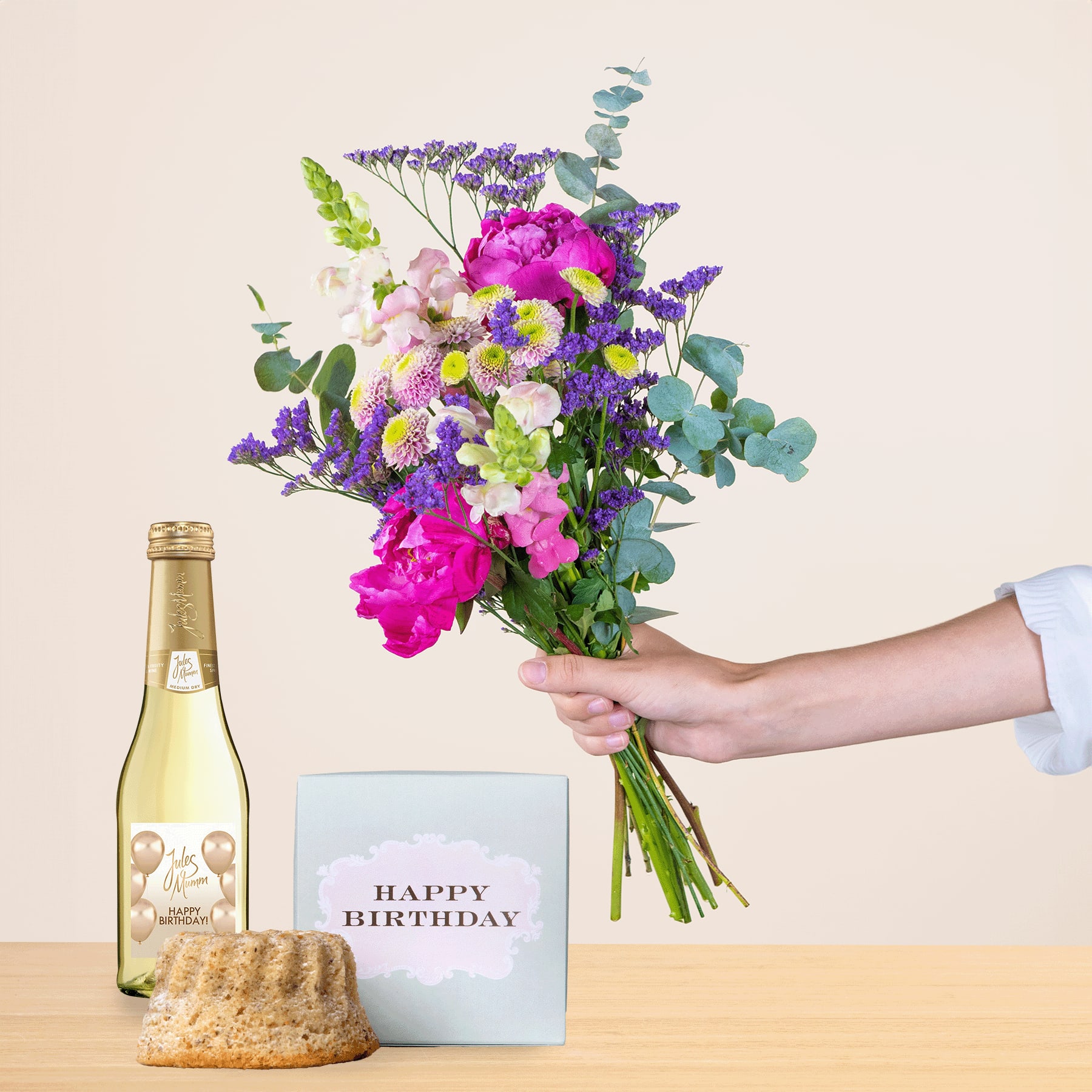 Ein leuchtender Strauß bunter Blumen, darunter rosa Pfingstrosen, lila Lavendel und weiße Gänseblümchen, wird zusammen mit einer Flasche Champagner und einer Geburtstagskarte auf einer Holzoberfläche in der Hand einer Person gehalten.
