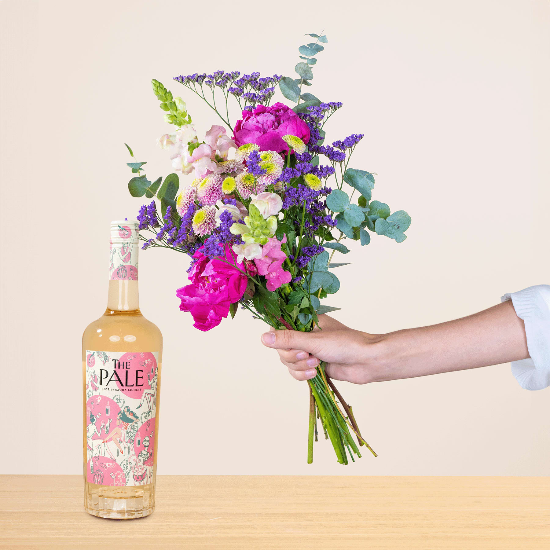Ein leuchtender Strauß bunter Blumen, darunter rosa Pfingstrosen, lila Lavendel und weiße Gänseblümchen, wird neben einer Flasche Pale Ale auf einer Holzoberfläche in der Hand gehalten.