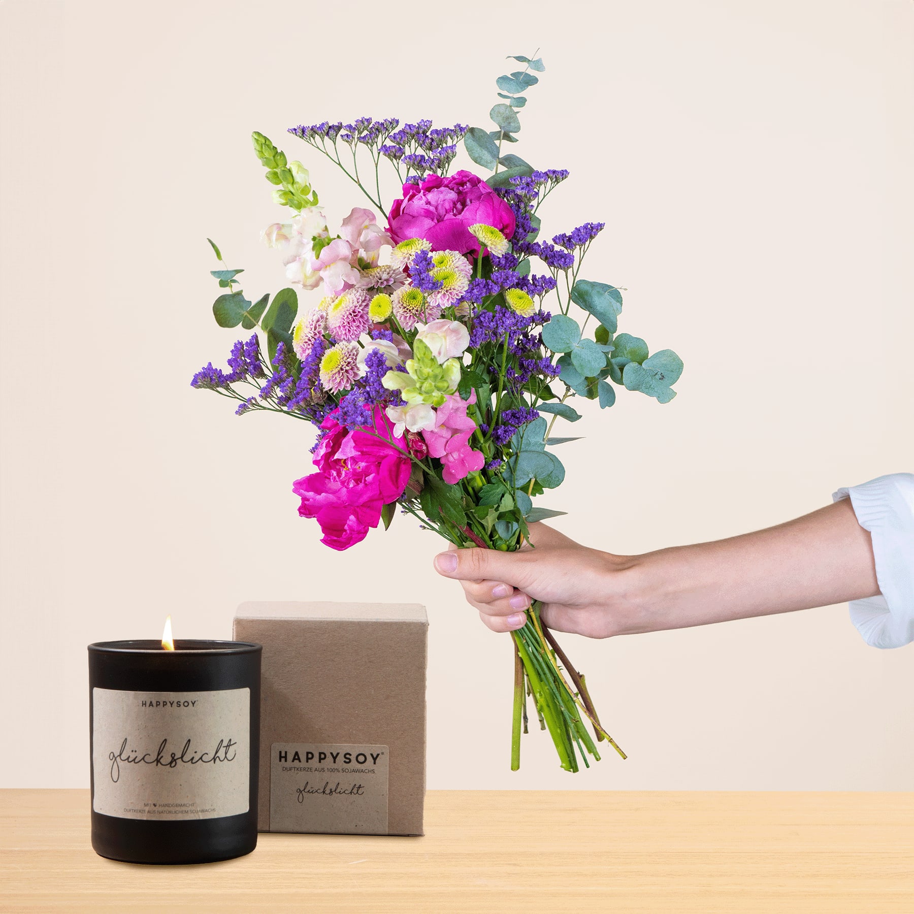 Eine Person hält einen leuchtenden Strauß bunter Blumen, darunter rosa Pfingstrosen, lila Lavendel und weiße Gänseblümchen, in der Hand, im Hintergrund sind eine Kerze und eine Geschenkbox zu sehen.