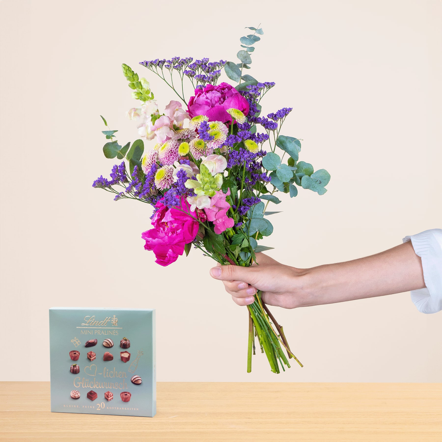 Ein leuchtender Strauß bunter Blumen, darunter rosa Pfingstrosen, lila Lavendel und weiße Gänseblümchen, wird vor einem schlichten Hintergrund in der Hand einer Person gehalten.