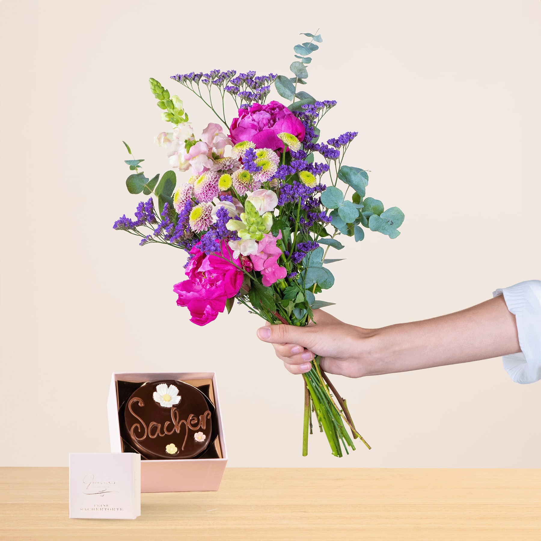 Eine Person hält einen leuchtend bunten Blumenstrauß, darunter rosa Pfingstrosen, lila Lavendel und grüne Blätter, in der Hand. Im Hintergrund liegen auf dem Tisch eine kleine Pralinenschachtel und eine Grußkarte.