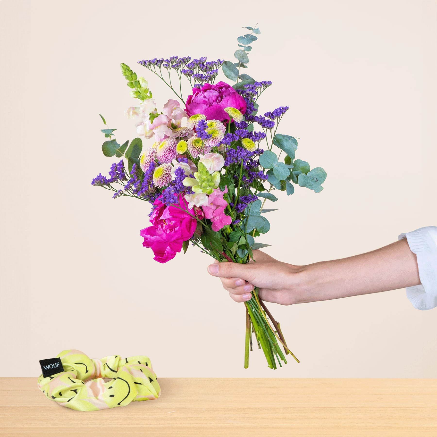 Ein leuchtender Strauß bunter Blumen, darunter rosa Pfingstrosen, lila Lavendel und grünes Laub, wird vor einem schlichten beigen Hintergrund in der Hand gehalten, im Vordergrund ist ein Stapel grüner Äpfel zu sehen.