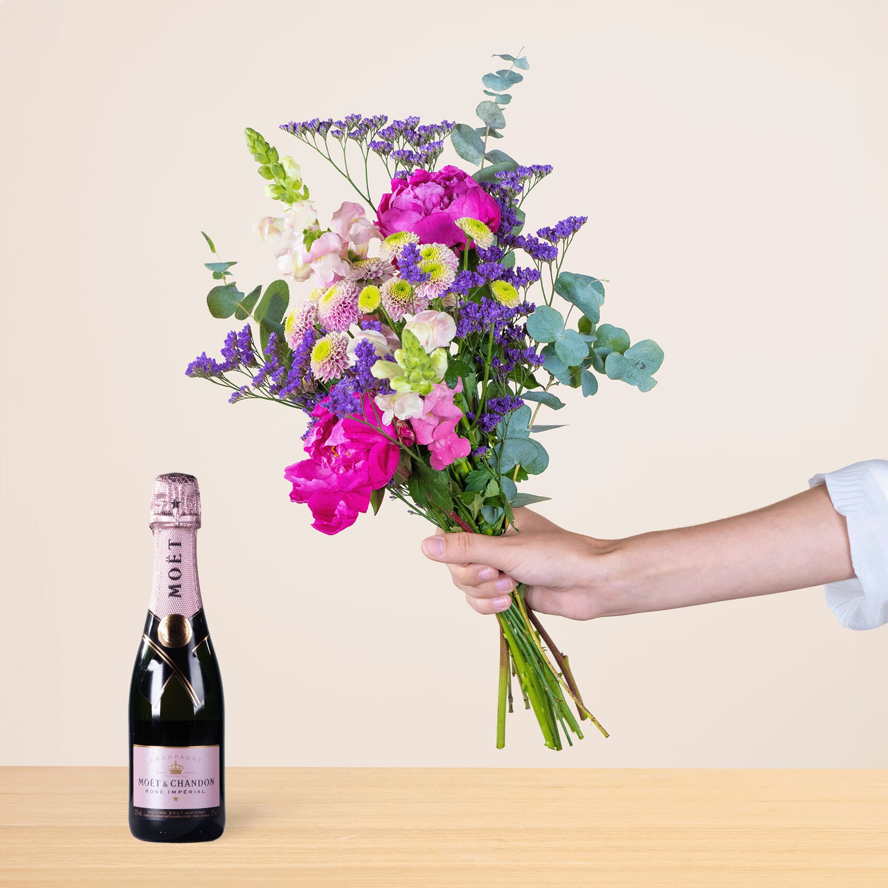 Ein leuchtender Strauß bunter Blumen, darunter rosa Pfingstrosen, lila Lavendel und grünes Laub, wird neben einer Flasche Champagner auf einer Holzoberfläche in der Hand einer Person gehalten.