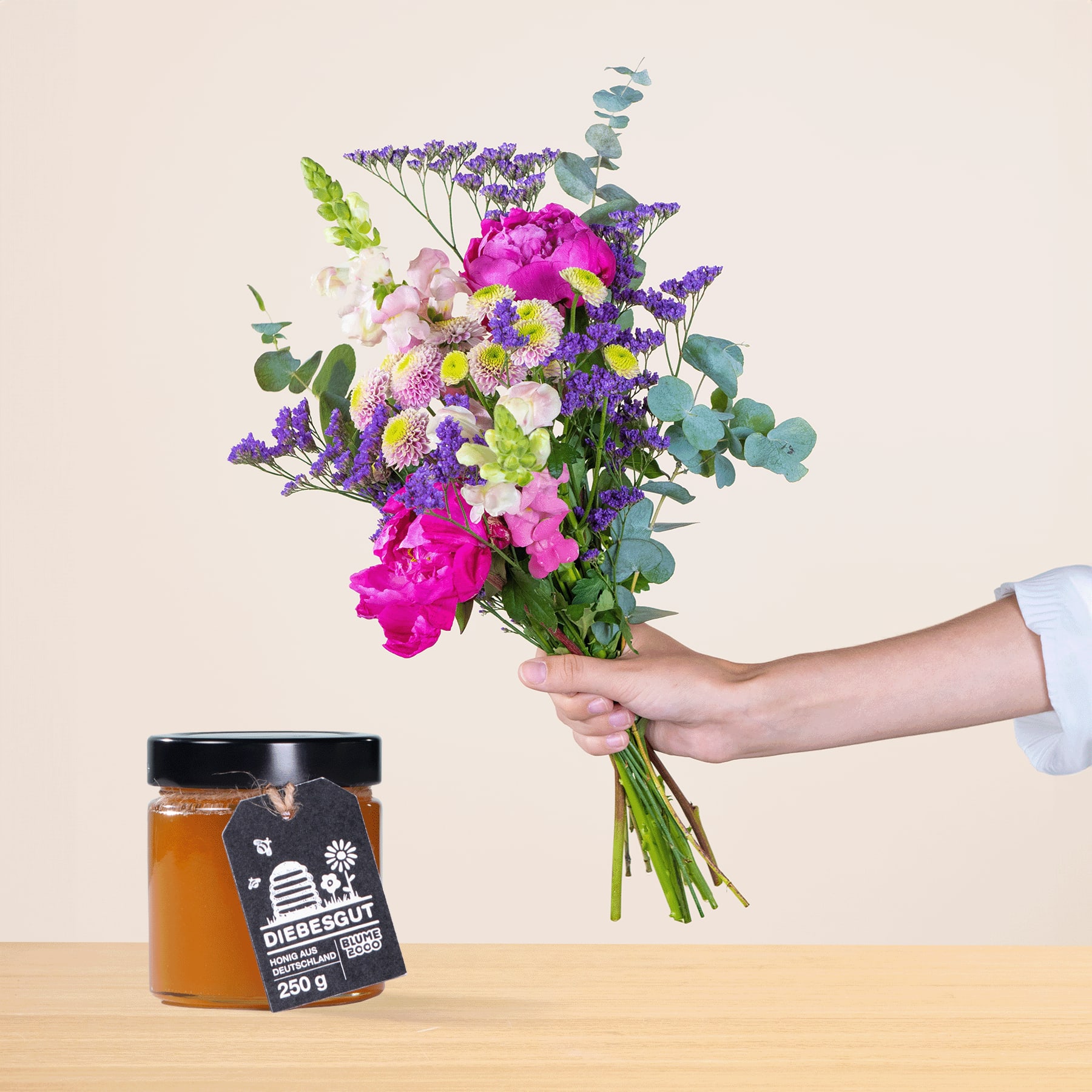 Eine Person hält einen leuchtend bunten Blumenstrauß, darunter rosa Pfingstrosen, lila Lavendel und grünes Laub, in der Hand. Im Vordergrund ist auf einer Holzoberfläche ein Glas Honig zu sehen.