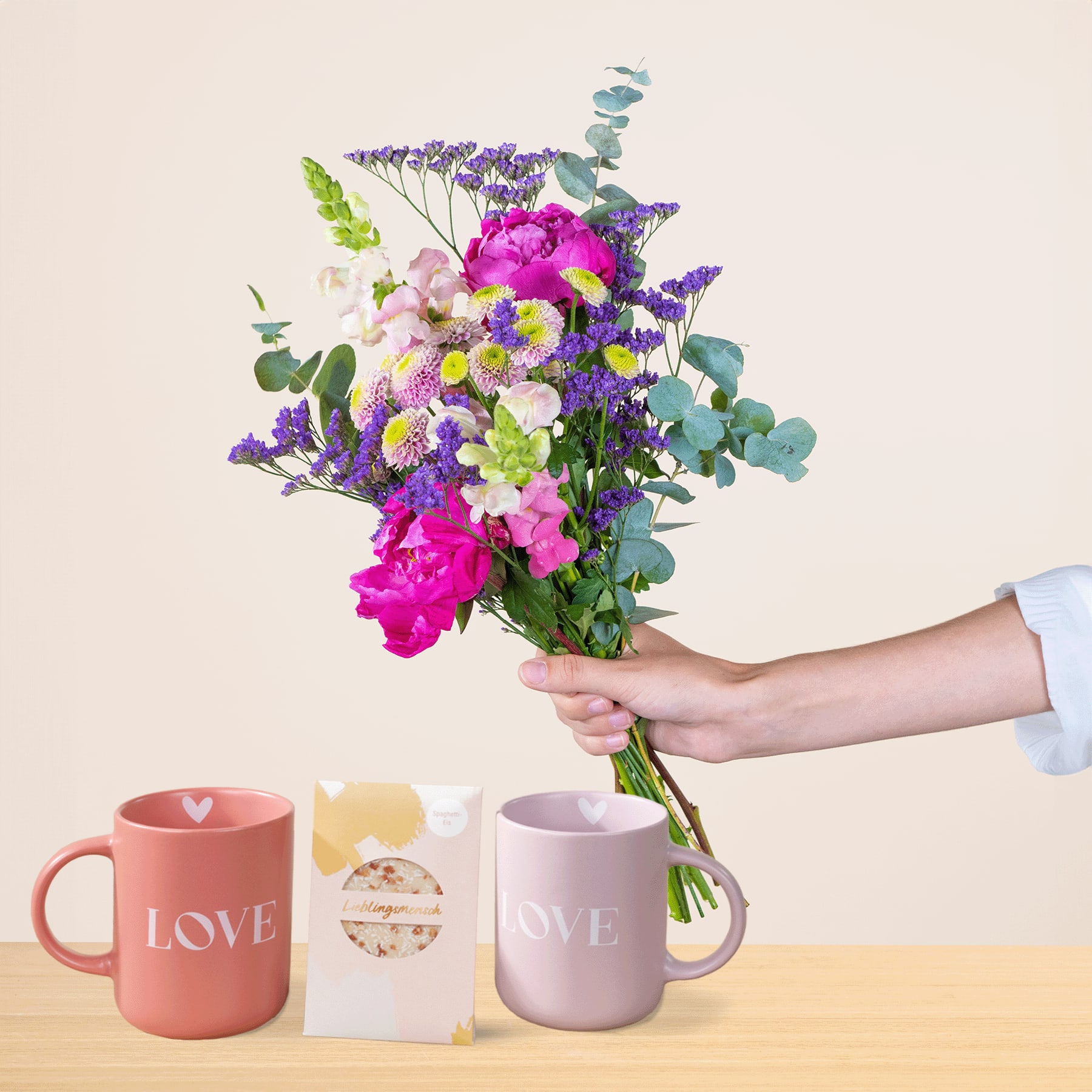 Eine Person hält einen leuchtend bunten Blumenstrauß, darunter rosa Pfingstrosen und lila Lavendel, in der Hand, begleitet von zwei Tassen mit dem Aufdruck „LOVE“ und einer kleinen Geschenkbox auf schlichtem Hintergrund.