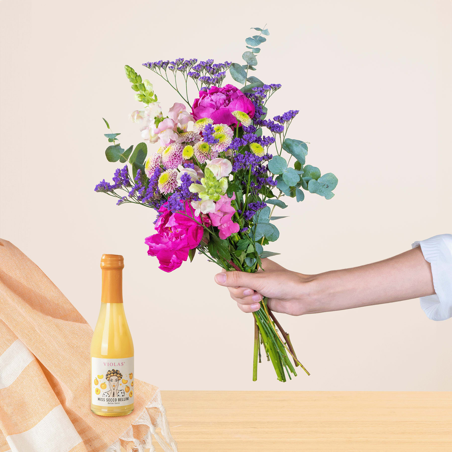 Ein leuchtender Strauß bunter Blumen, darunter rosa Pfingstrosen, violette Schwertlilien und Grünpflanzen, wird vor einem schlichten Hintergrund in den Händen einer Person gehalten, auf der eine Flasche Wein zu sehen ist.