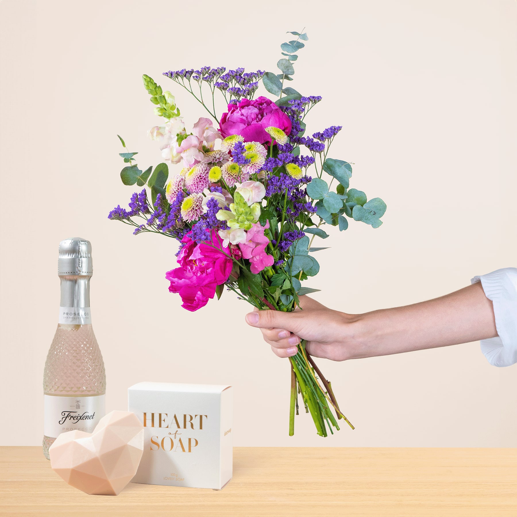 Eine Person hält einen leuchtend bunten Blumenstrauß, darunter rosa Pfingstrosen, violette Schwertlilien und grüne Blätter, in der Hand. Vor einem schlichten Hintergrund stehen eine Parfümflasche und ein Stück Seife.