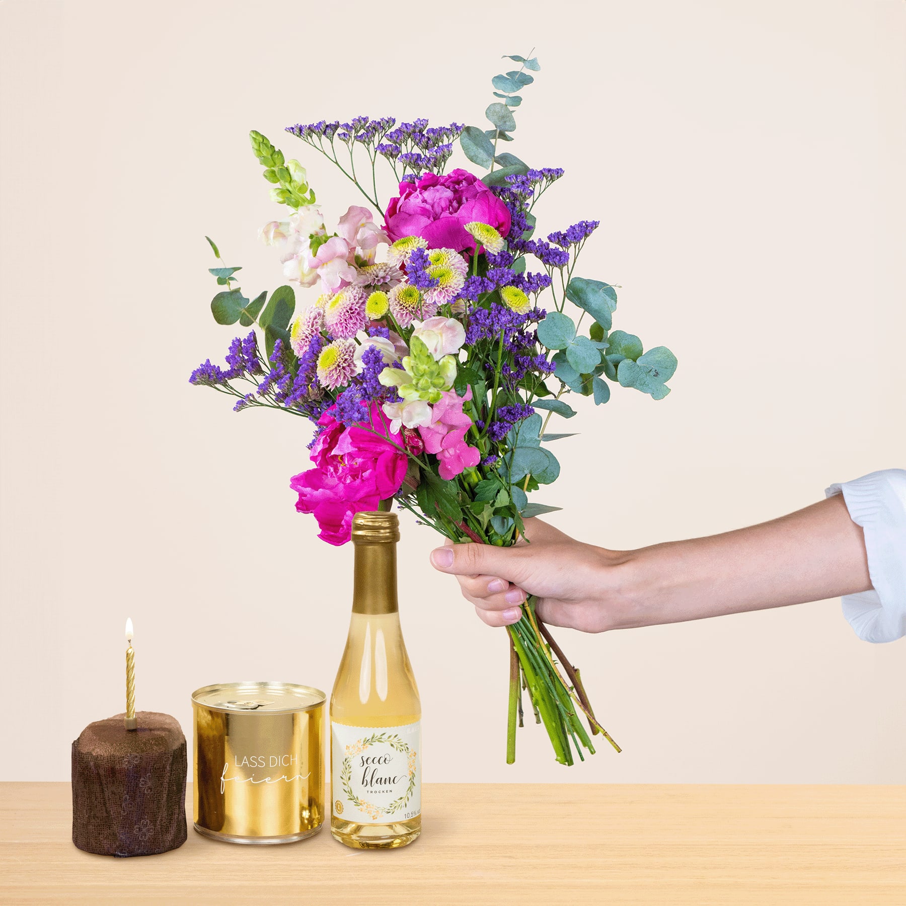 Ein leuchtender Strauß bunter Blumen, darunter rosa Pfingstrosen, lila Lavendel und gelbe Gänseblümchen, wird in der Hand gehalten, vor einem schlichten Hintergrund mit dekorativen Kerzen und Gefäßen.