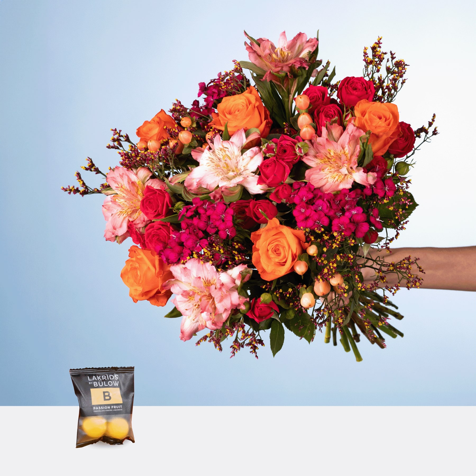 Ein farbenfroher Strauß roter, orangefarbener und rosa Blumen, darunter Rosen und Dahlien, wird vor einem schlichten blauen Hintergrund in den Händen einer Person gehalten. Im Vordergrund steht eine kleine Tüte Zitronen.