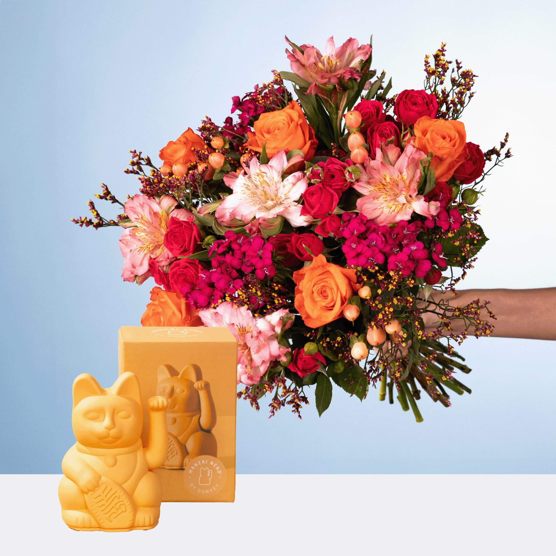 Ein leuchtender Strauß roter, orangefarbener und rosa Blumen, darunter Rosen und Dahlien, wird neben einer gelben Keramikfigur vor einem blauen Hintergrund gehalten.