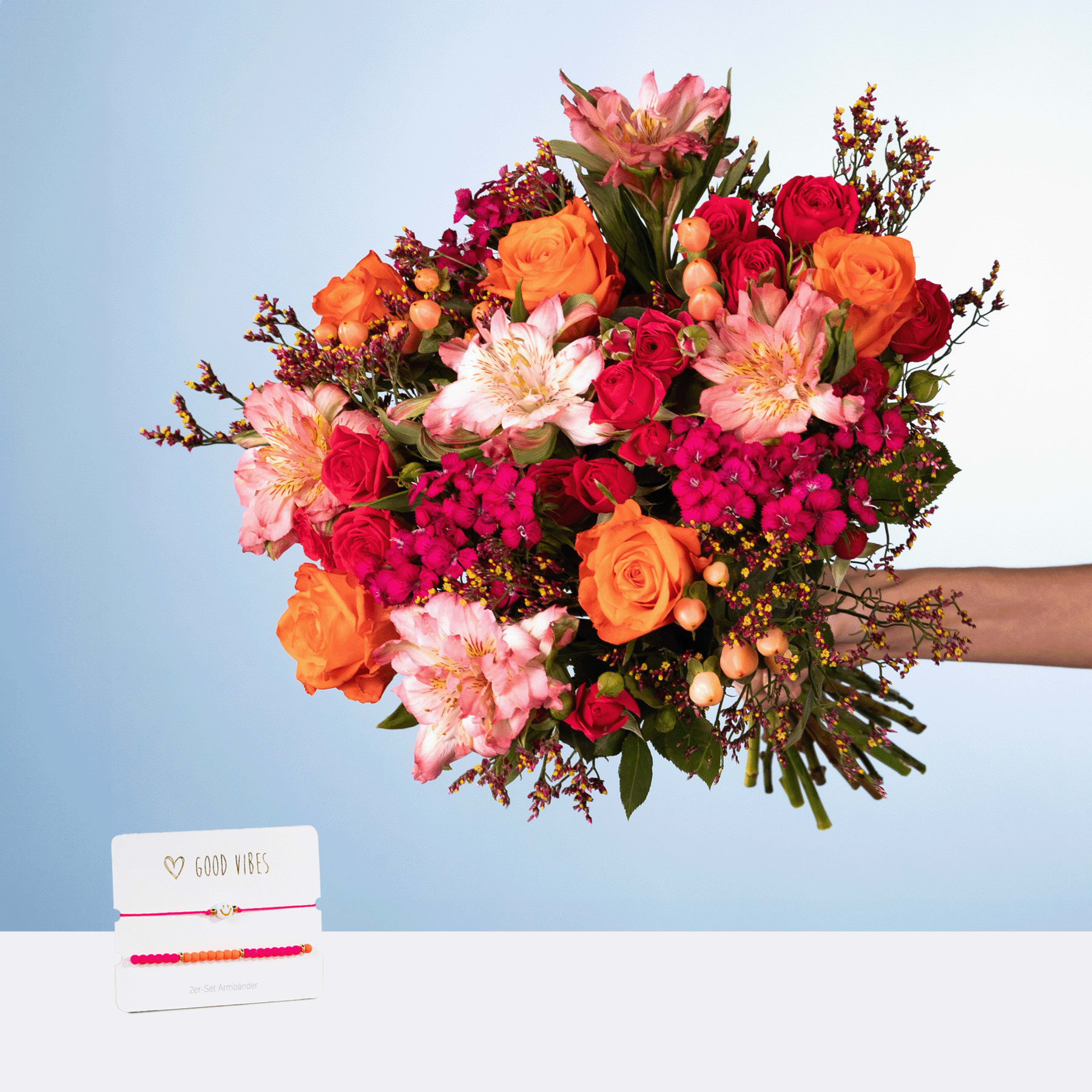 Ein farbenfroher Strauß roter, orangefarbener und rosa Blumen, darunter Rosen, Dahlien und andere Blüten, wird vor einem schlichten blauen Hintergrund in den Händen einer Person gehalten.