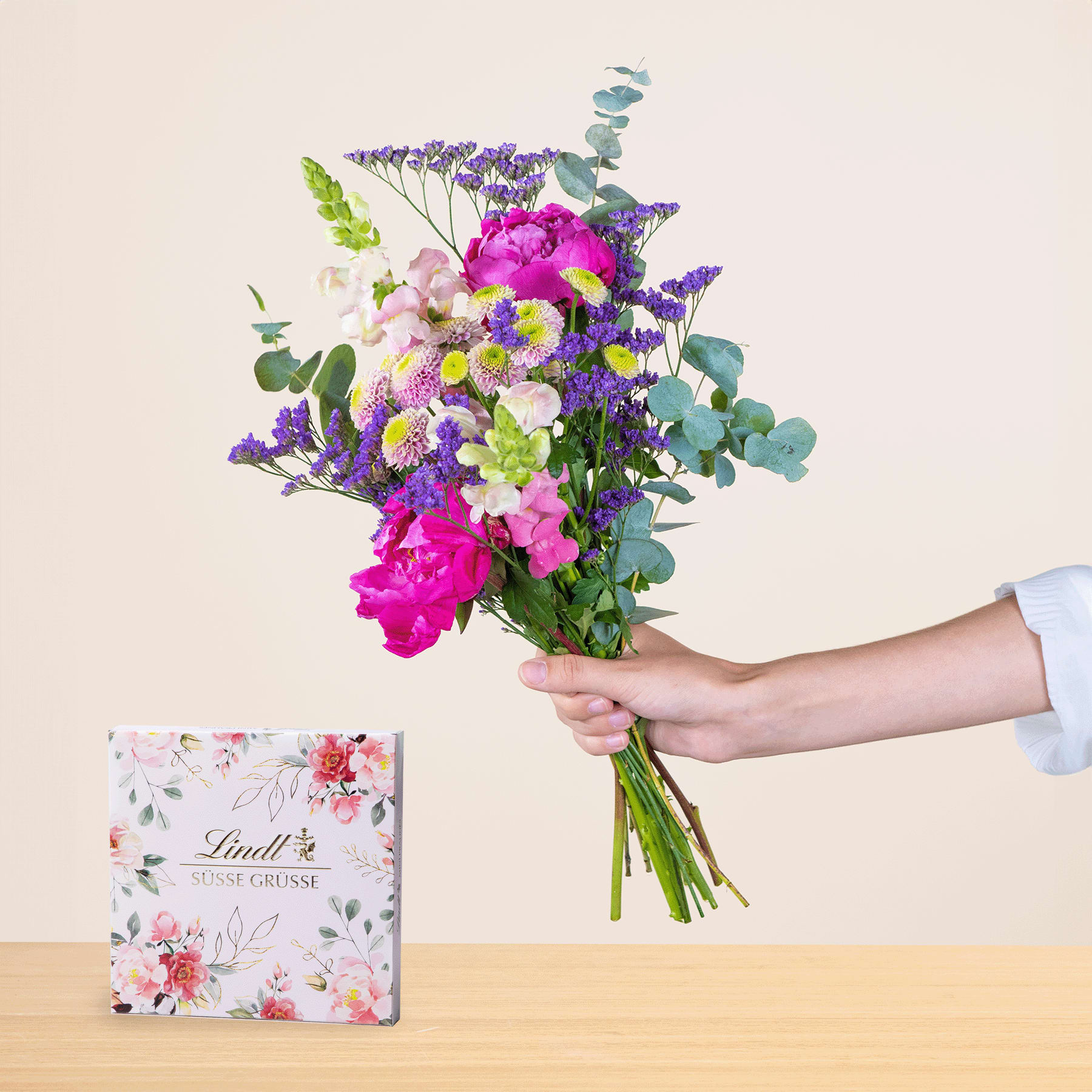Eine Person hält einen leuchtend bunten Blumenstrauß, darunter rosa Pfingstrosen, lila Lavendel und weiße Gänseblümchen, in der Hand. Im Hintergrund ist eine Geschenkbox mit Blumenmuster zu sehen.