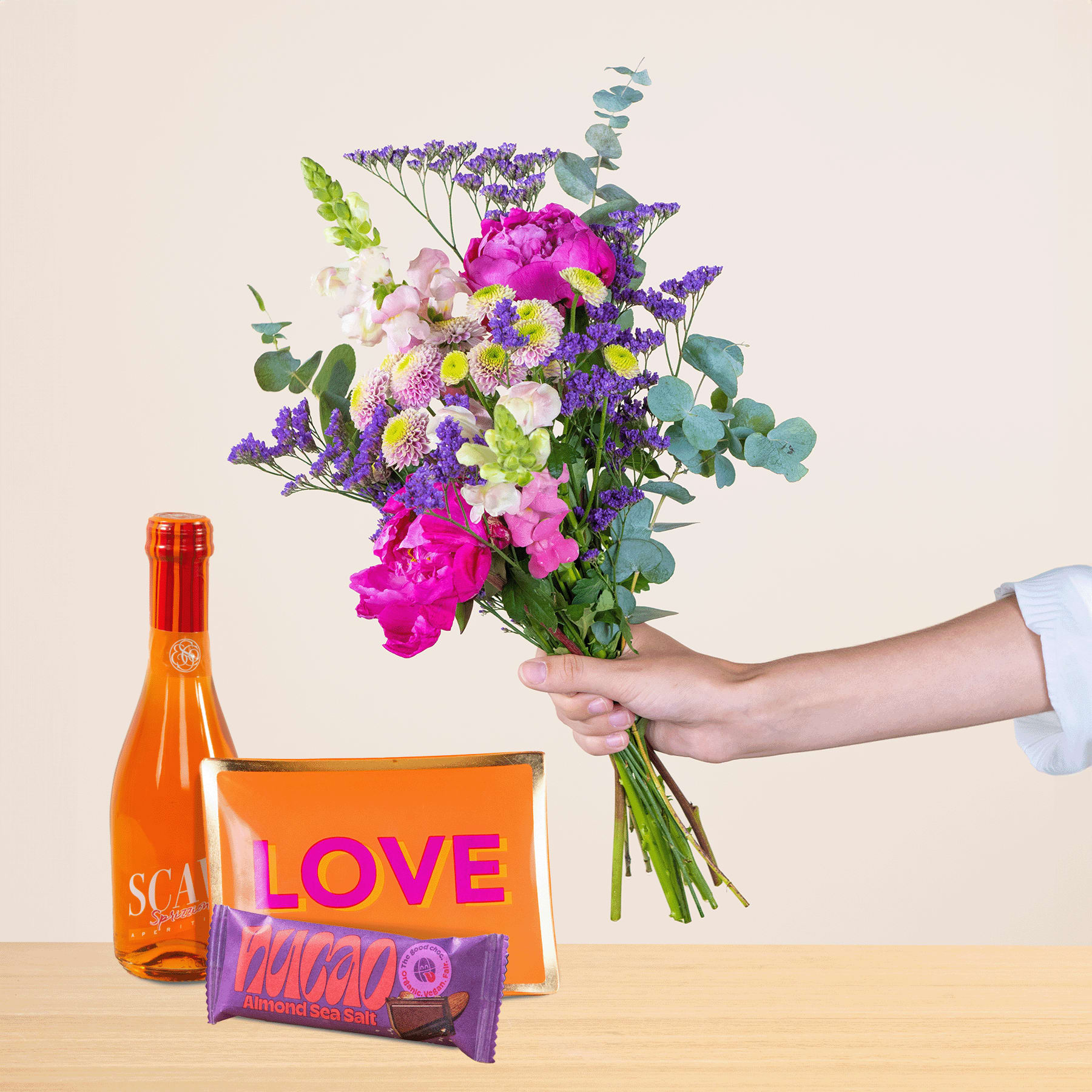 Eine Person hält einen leuchtend bunten Blumenstrauß, darunter rosa Pfingstrosen und lila Lavendel, in der Hand, begleitet von einer Flasche Orangenlimonade und einer herzförmigen Pralinenschachtel auf einer Holzoberfläche.