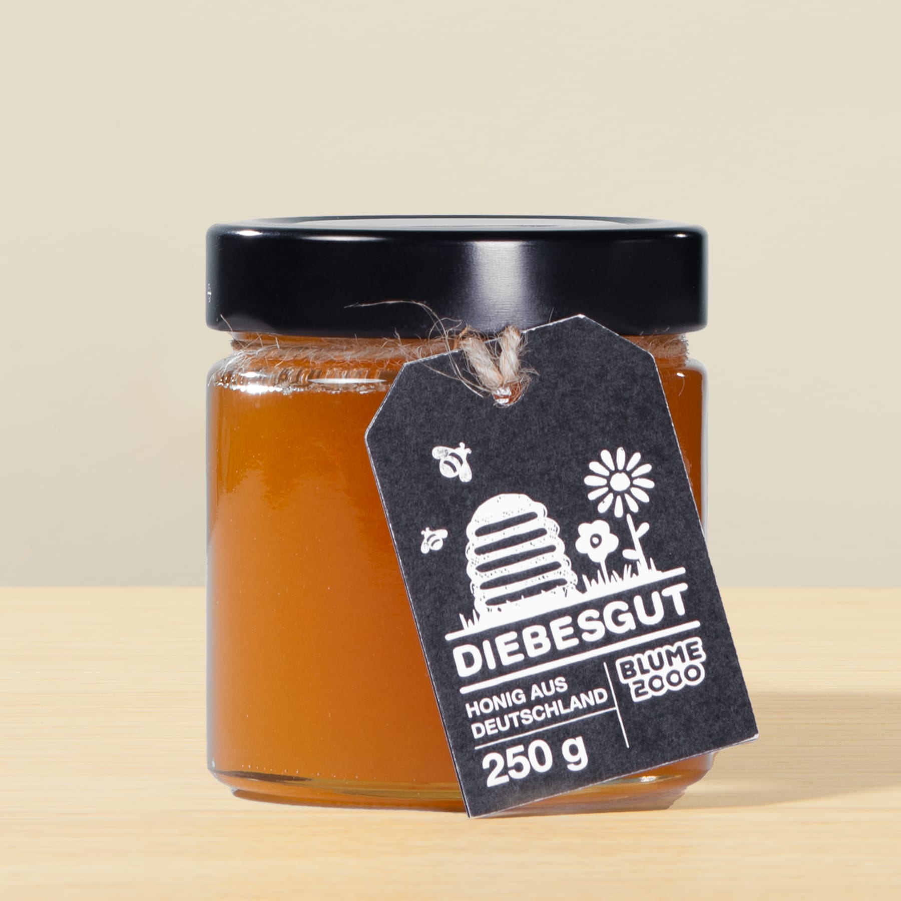 Ein mit goldenem Honig gefülltes Glas mit einem schwarzen Etikett mit Logo und Text steht auf einer Holzoberfläche vor einem hellen Hintergrund.