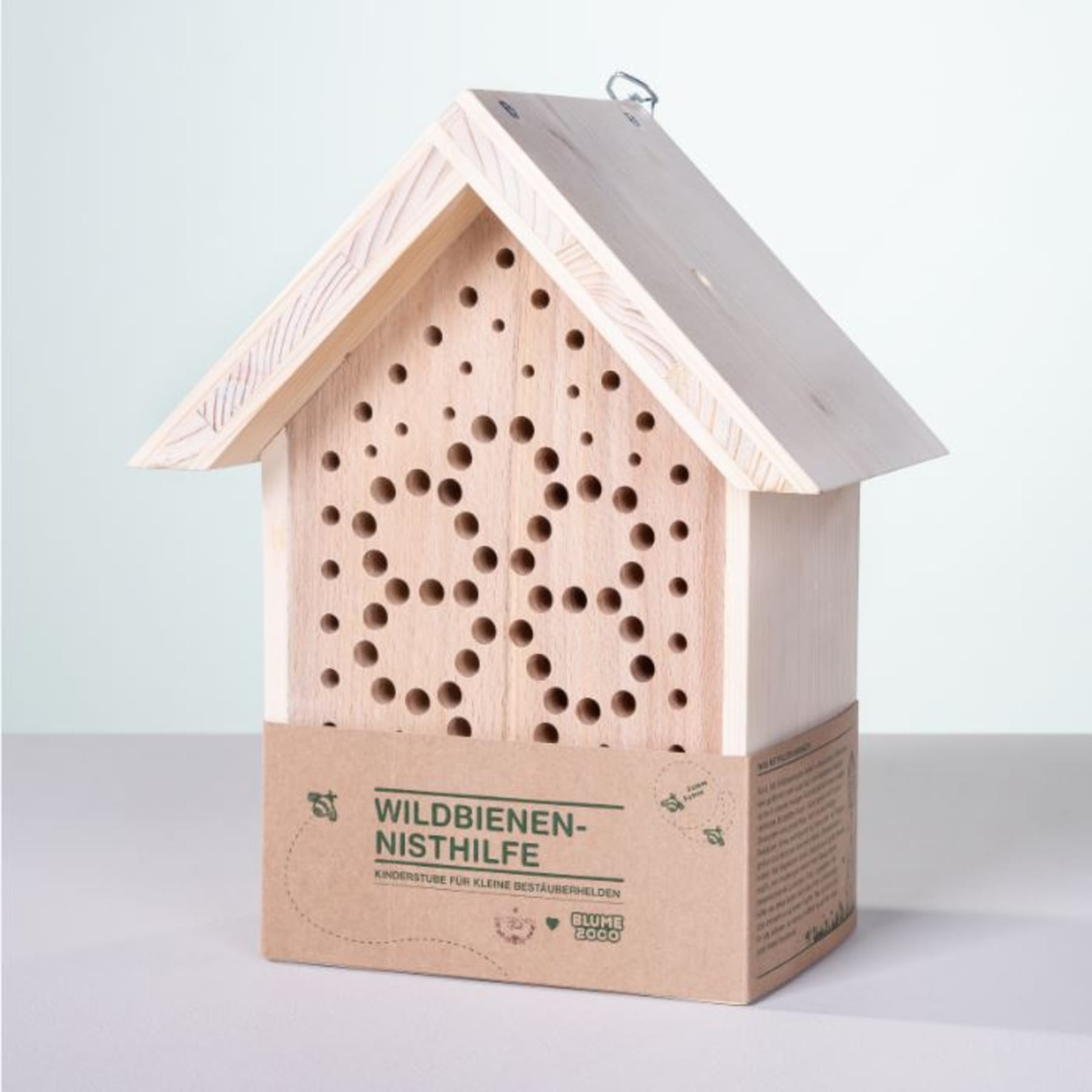 Ein Insektenhotel aus Holz mit schrägem Dach und zahlreichen Löchern, die Insekten als Unterschlupf dienen, platziert auf einer grauen Fläche vor hellem Hintergrund.