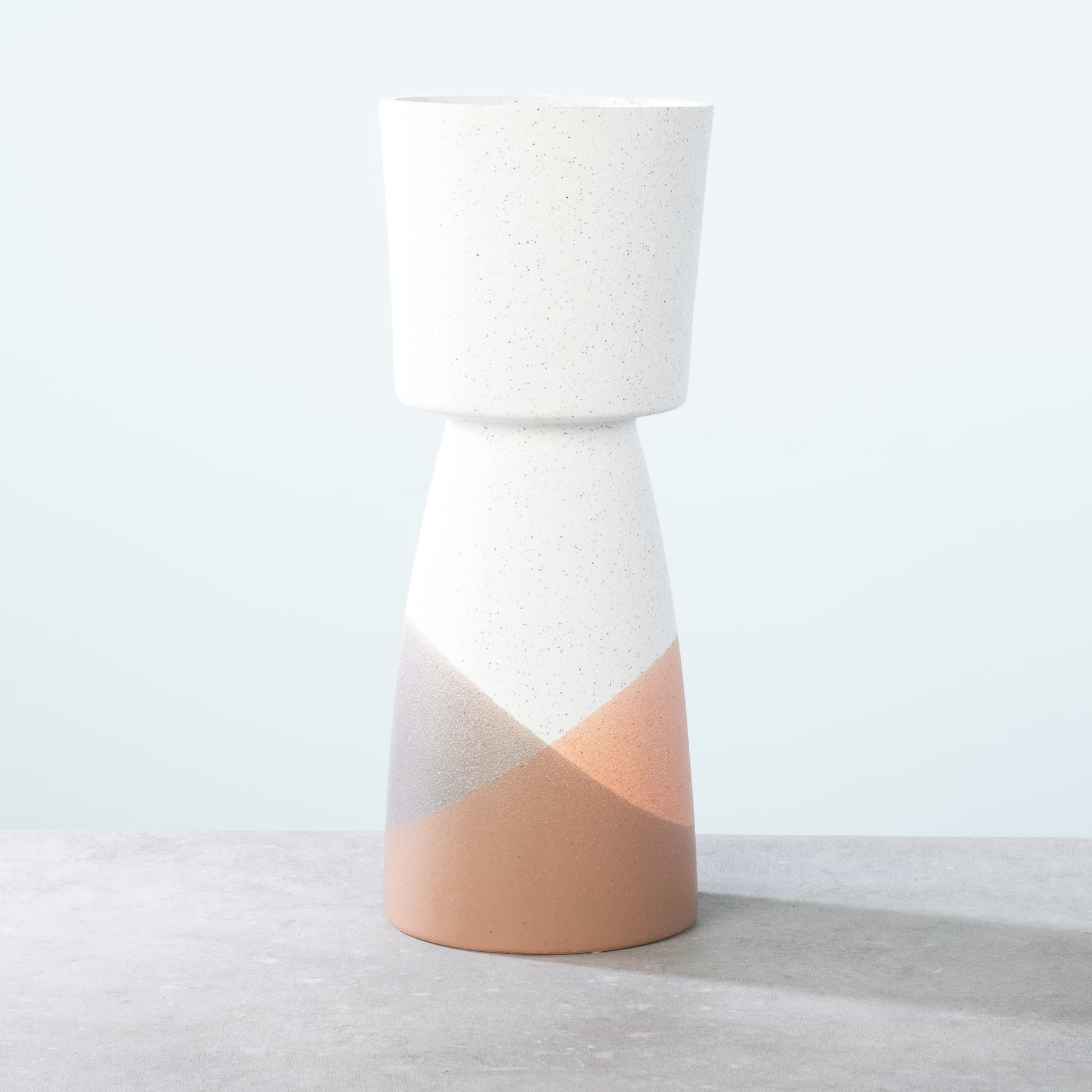 Eine weiße Keramikvase mit zweifarbigem Design, bestehend aus einer hellpfirsichfarbenen unteren Hälfte und einer weißen oberen Hälfte, die auf einer hellen Oberfläche vor einem schlichten weißen Hintergrund ruht.