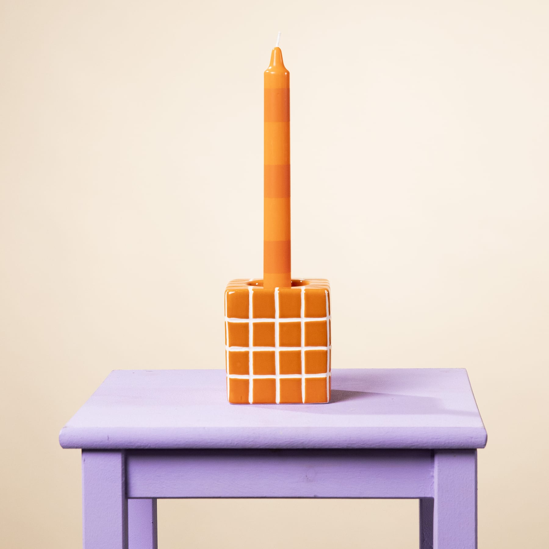 Eine leuchtend orangefarbene Kerze steht auf einem lavendelfarbenen Holztisch vor einem schlichten, hellen Hintergrund.