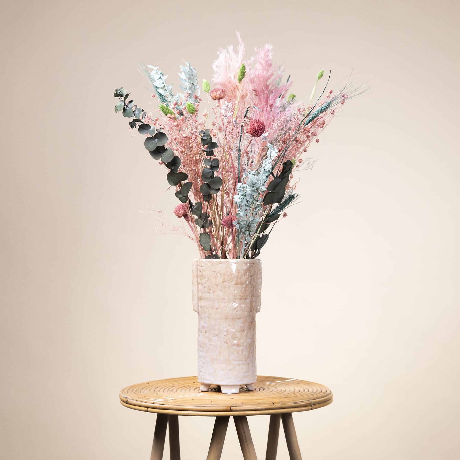 Eine Vase mit einem farbenfrohen Strauß getrockneter Blumen in Rosa, Weiß und Lila steht auf einem Holzsockel vor einem neutralen beigen Hintergrund.