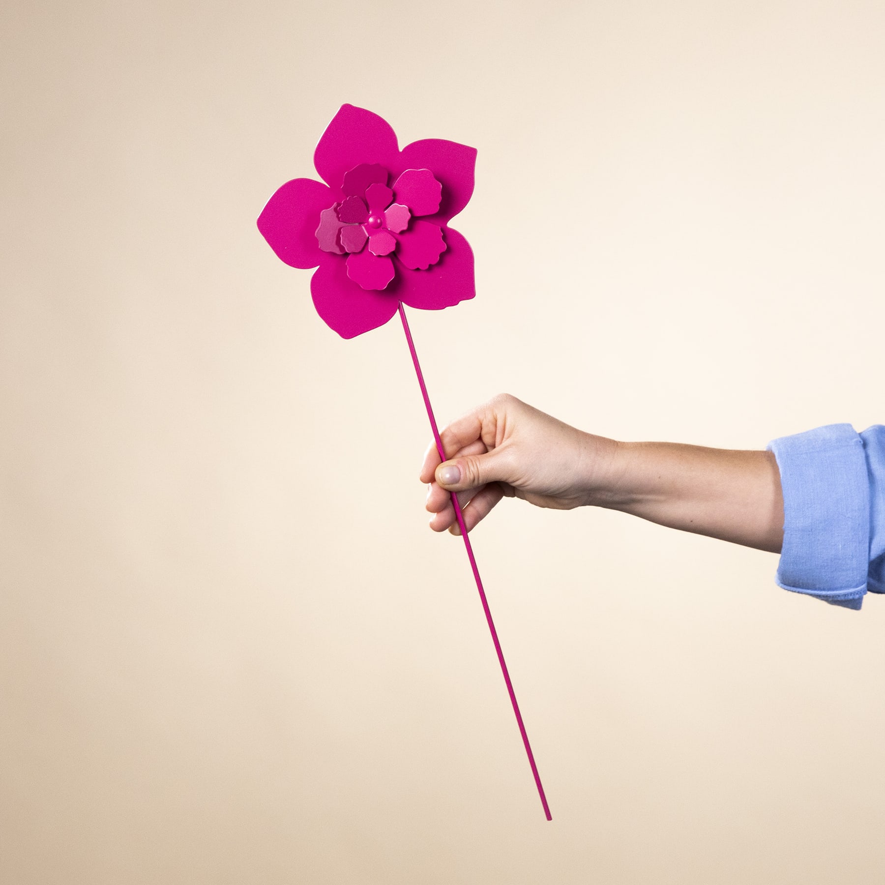 Eine leuchtend fuchsiafarbene Papierblume wird vor einem neutralen, beigen Hintergrund in der Hand gehalten.