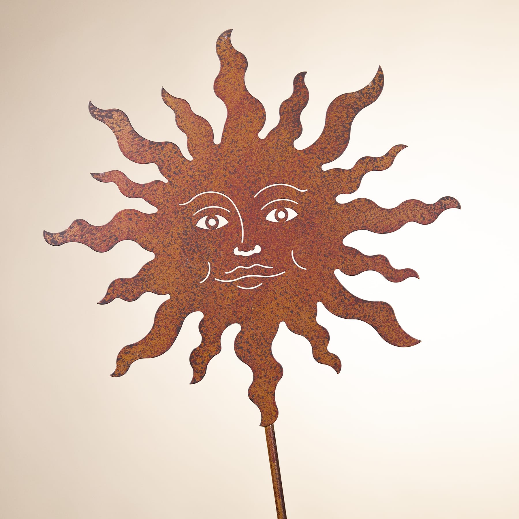 Eine rustikale, sonnenförmige Metallskulptur mit einem Gesicht und von ihr ausgehenden Strahlen vor einem schlichten, hellen Hintergrund.