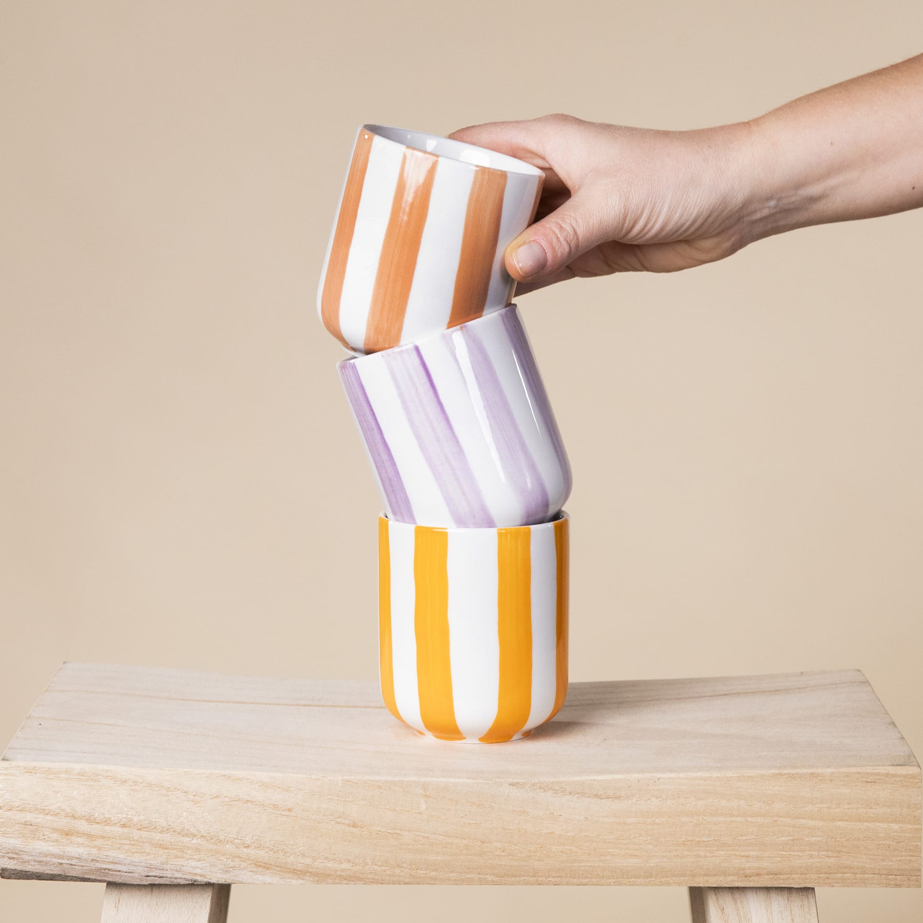 Eine Hand stapelt bunt gestreifte Keramiktassen oder -becher auf einer Holzoberfläche vor einem beigen Hintergrund.