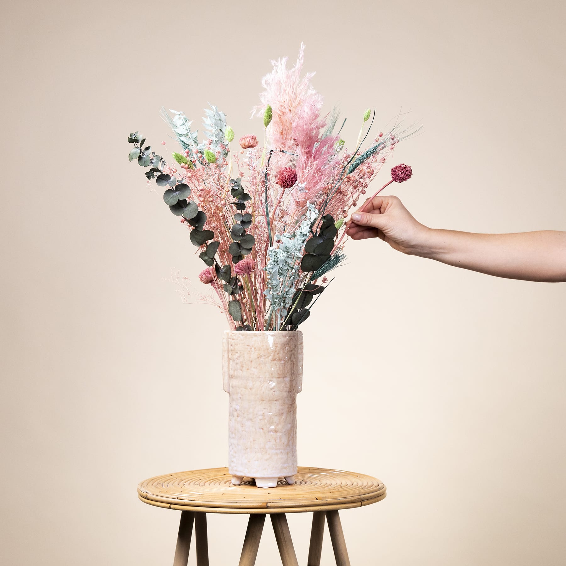 Eine Hand hält eine Vase mit einem leuchtenden Strauß bunter Trockenblumen und Pflanzen vor einem neutralen beigen Hintergrund.