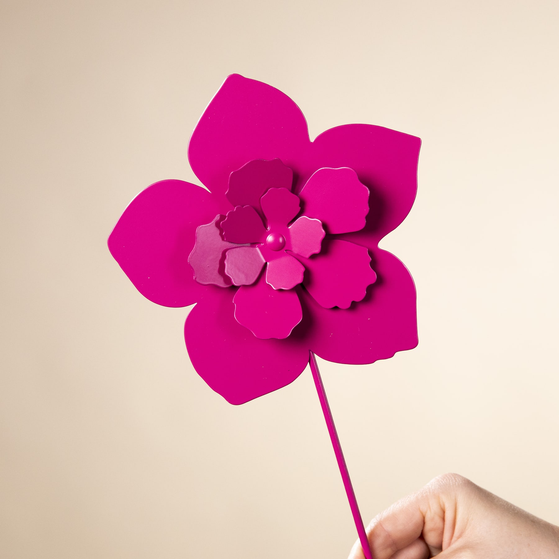 Eine leuchtende, fuchsiafarbene Papierblume mit mehrschichtigen Blütenblättern hebt sich von einem neutralen, beigen Hintergrund ab.