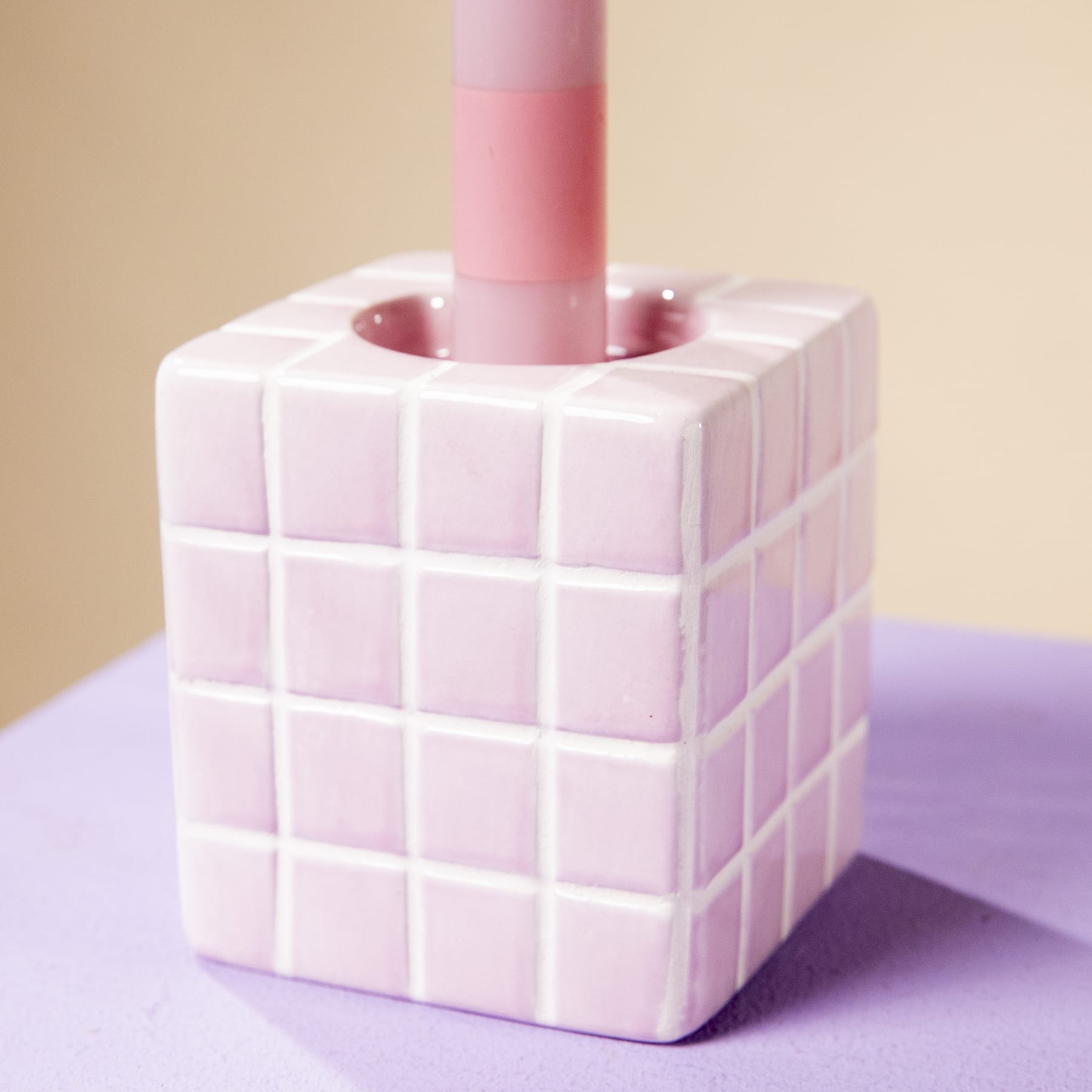 Ein weißes, würfelförmiges Objekt mit einer rosa Kerze, die aus der Mitte herausragt und auf einer violetten Oberfläche vor einem beigen Hintergrund ruht.