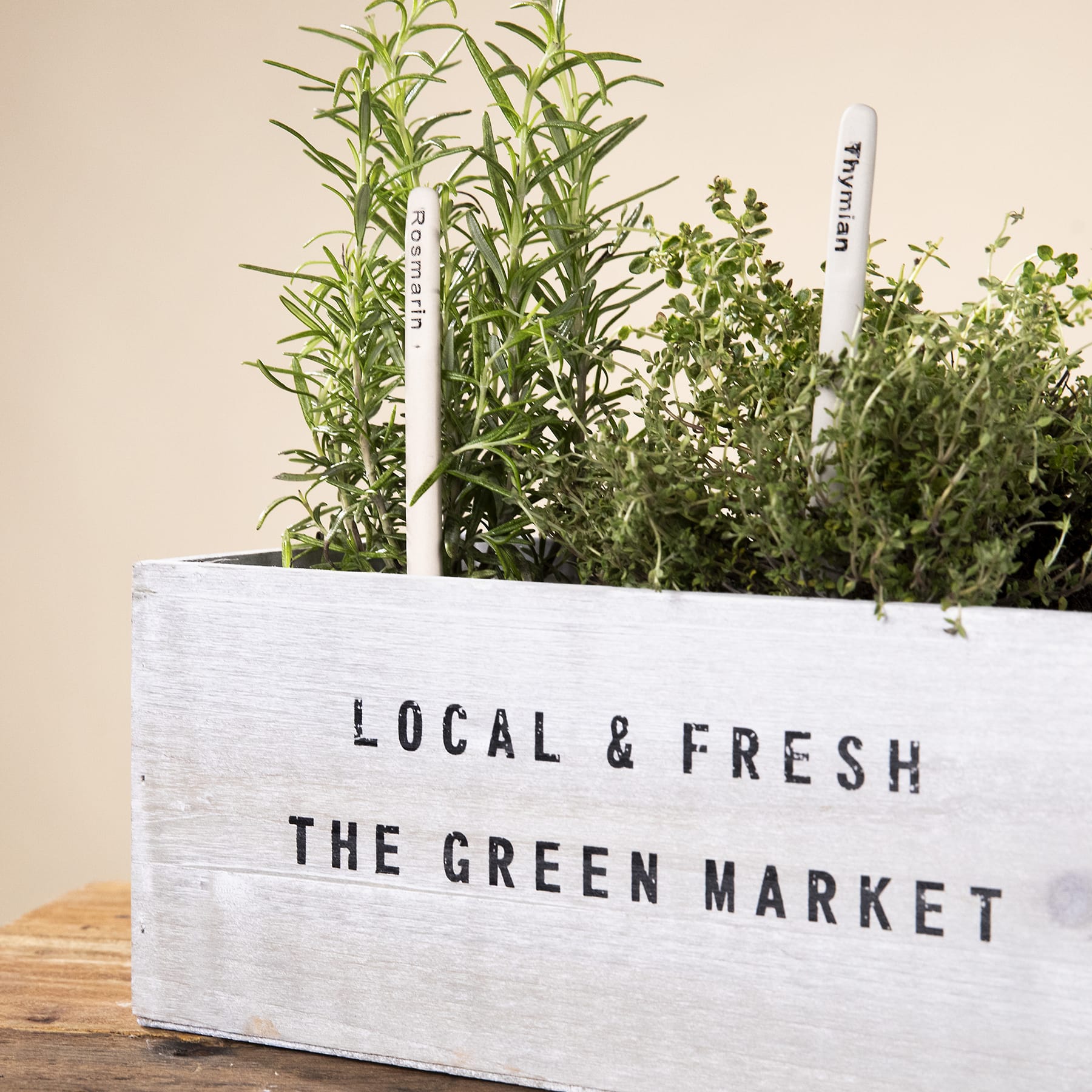 Eine Holzkiste gefüllt mit verschiedenen frischen Kräutern und Pflanzen, mit einem Etikett mit der Aufschrift „LOCAL & FRESH THE GREEN MARKET“, steht auf einer Holzoberfläche vor einem hellen Hintergrund.