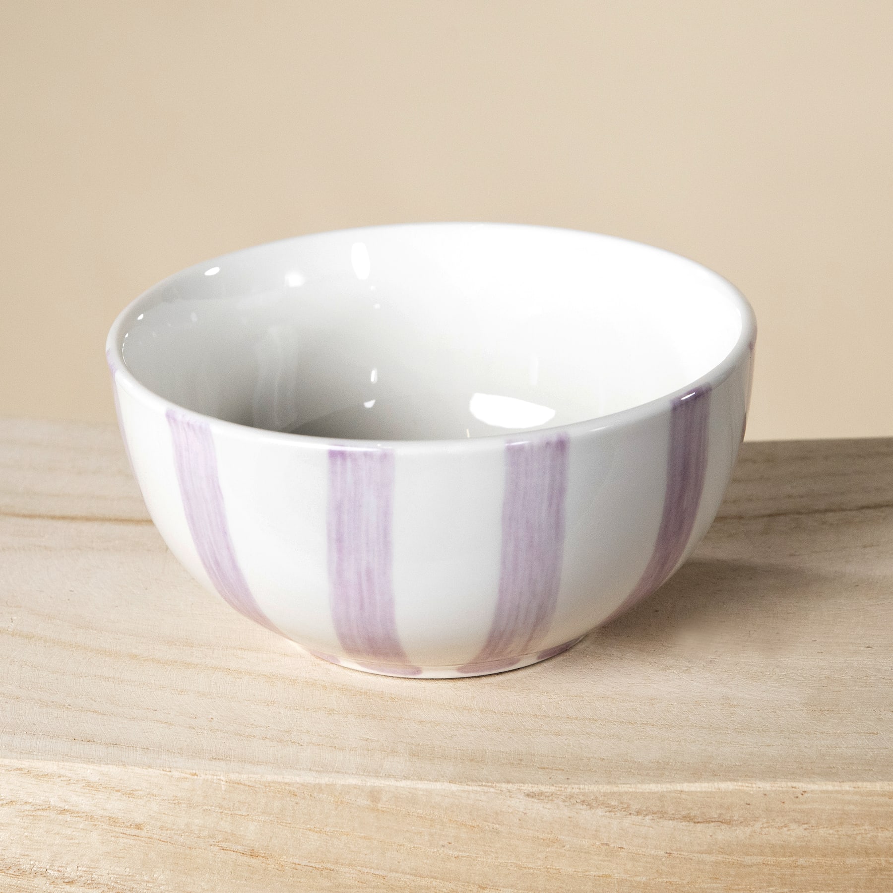 Eine weiße Keramikschale mit dezenten violetten Streifen steht auf einer Holzoberfläche vor einem beigen Hintergrund.