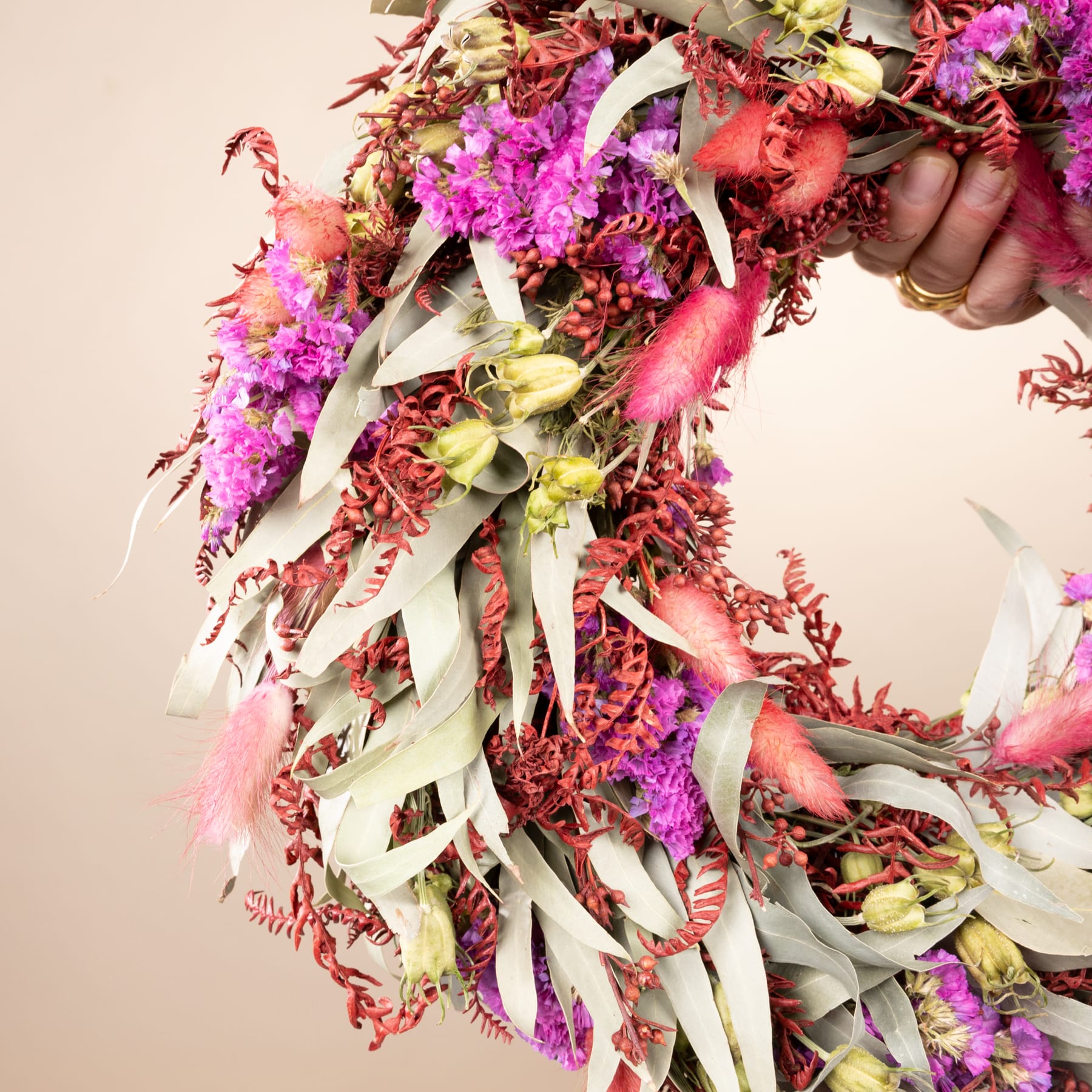 Jemand hält vor einem neutralen Hintergrund ein lebendiges und farbenfrohes Blumenarrangement mit einer Mischung aus rosa, roten und weißen Blumen sowie verschiedenen Grünpflanzen in den Händen.