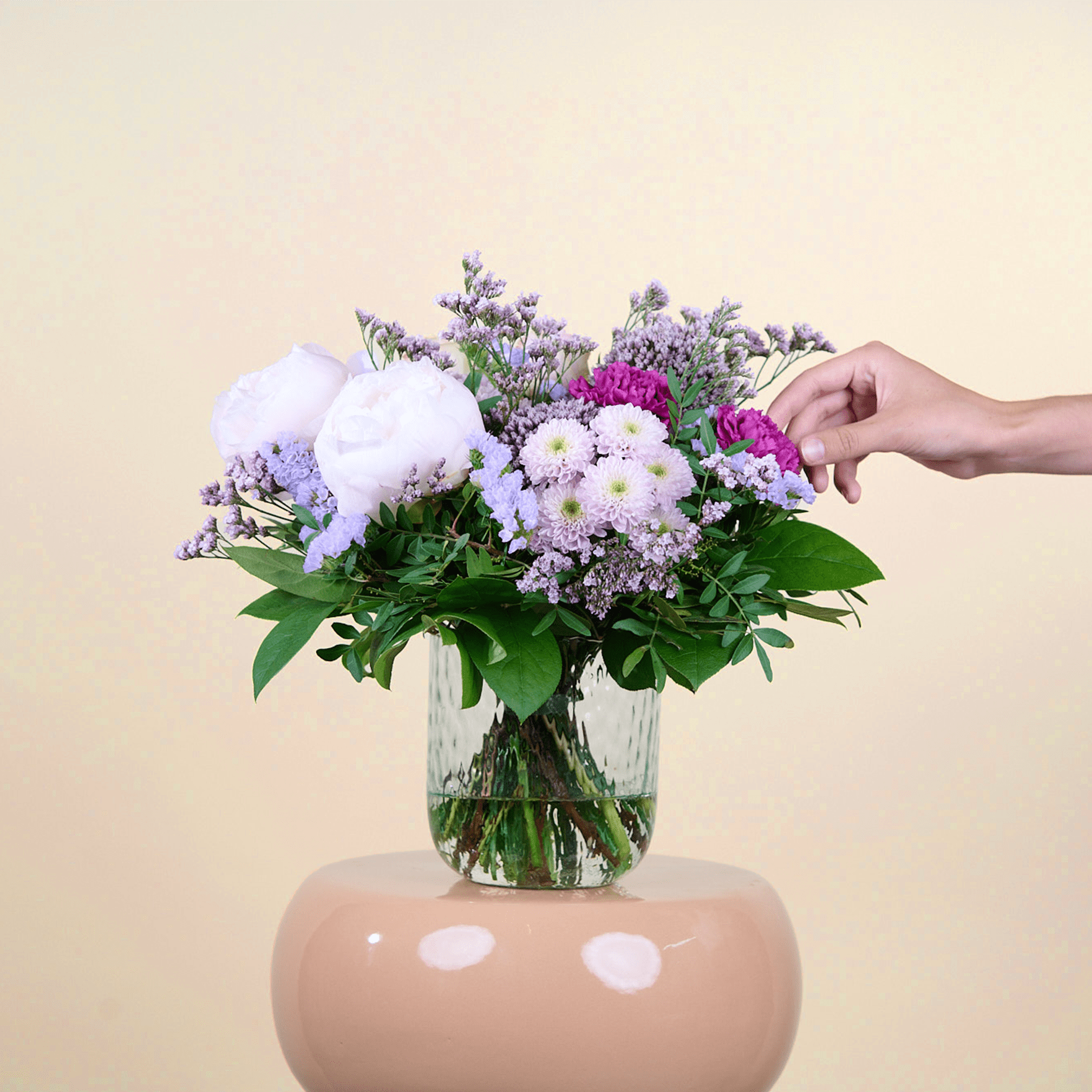 Ein Strauß leuchtend violetter, weißer und rosa Blumen in einer Glasvase. Eine Hand streckt sich hinein, um das Arrangement vor einem schlichten beigen Hintergrund zurechtzurücken.