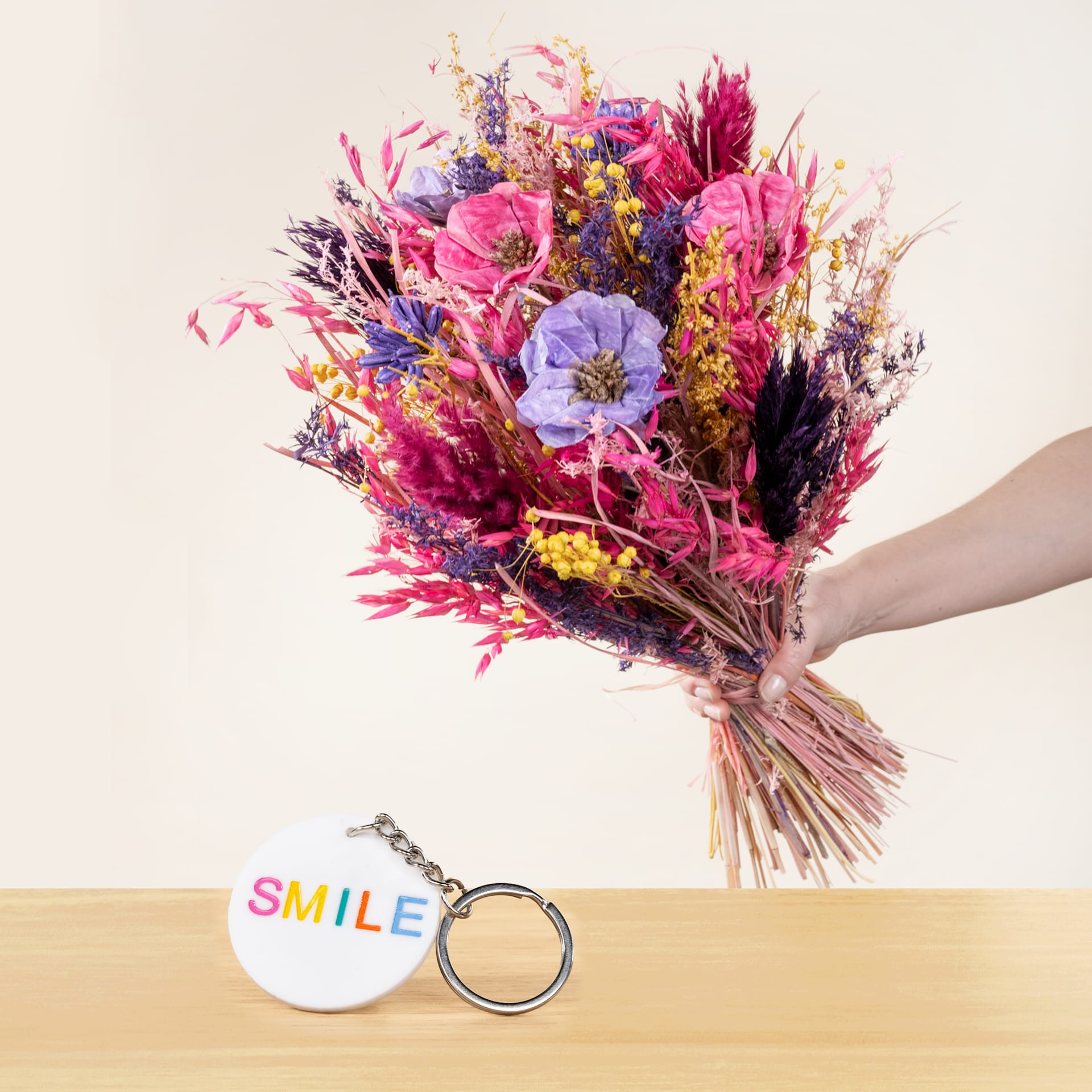 Eine Person hält einen leuchtend bunten Blumenstrauß, darunter rosa und violette Blüten, in der Hand und ruht auf einer Holzoberfläche neben einem kleinen runden Gegenstand, auf dem das Wort „SMILE“ steht.