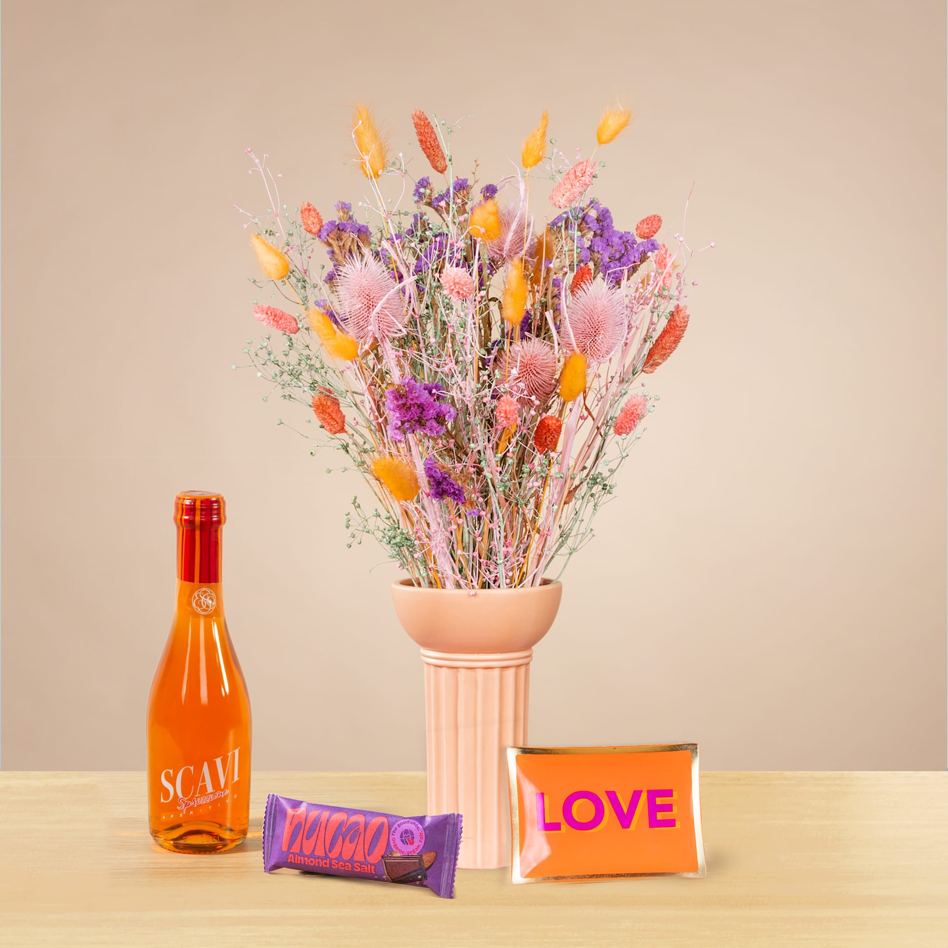 Ein leuchtender Strauß bunter Trockenblumen in einer Terrakotta-Vase steht auf einer Holzoberfläche, begleitet von einer Flasche Orangenflüssigkeit und zwei kleinen Dekorationsgegenständen.